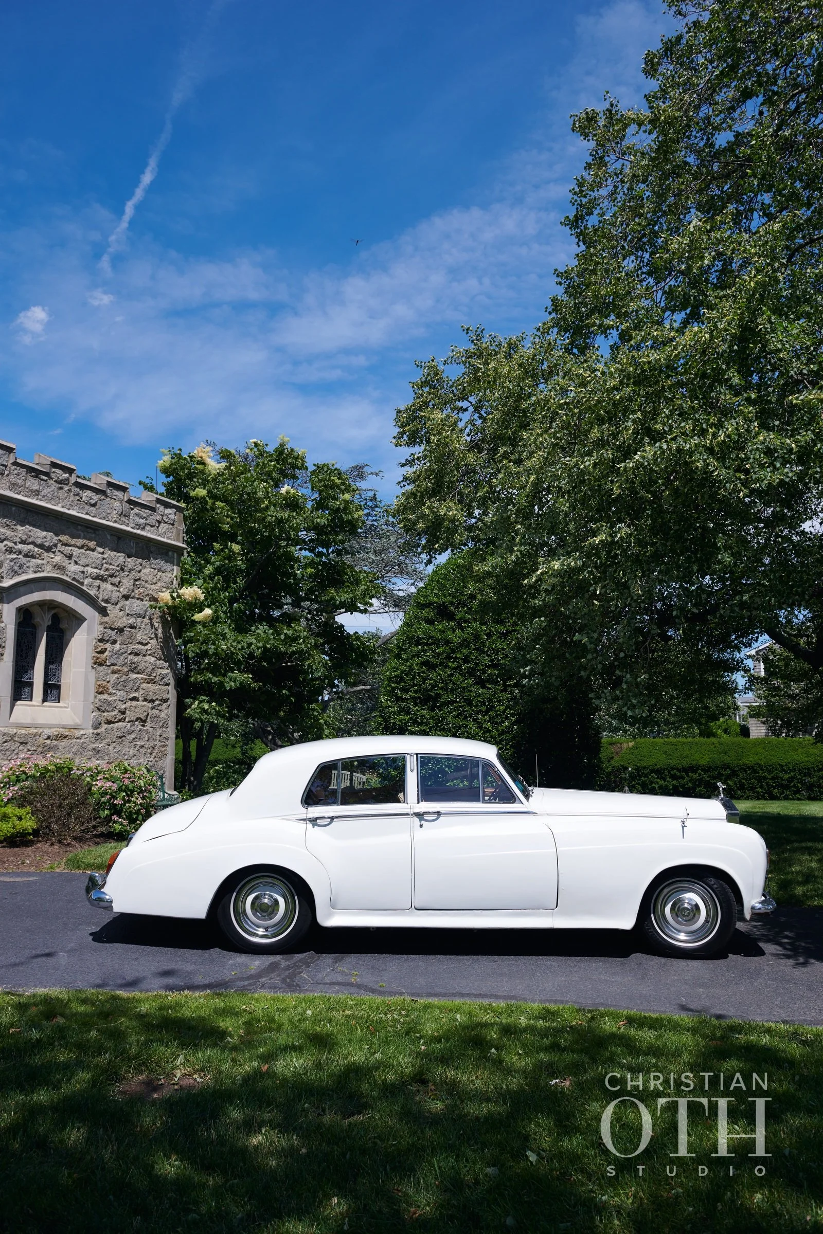 A vintage white luxury sedan parked on a paved driveway in front of a stone building with a window, surrounded by green trees and bushes, under a blue sky with some clouds.