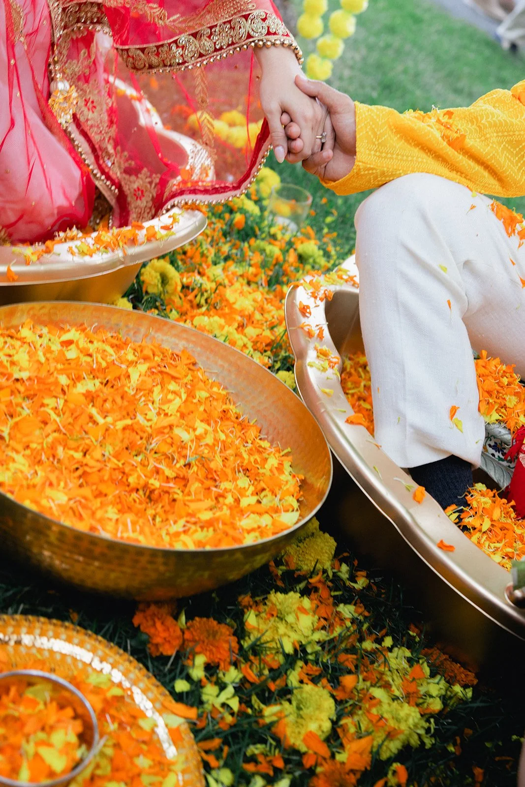 A close-up of a hand holding another hand during a traditional Indian wedding ceremony, with orange and yellow flower petals scattered around and in areca leaf plates.