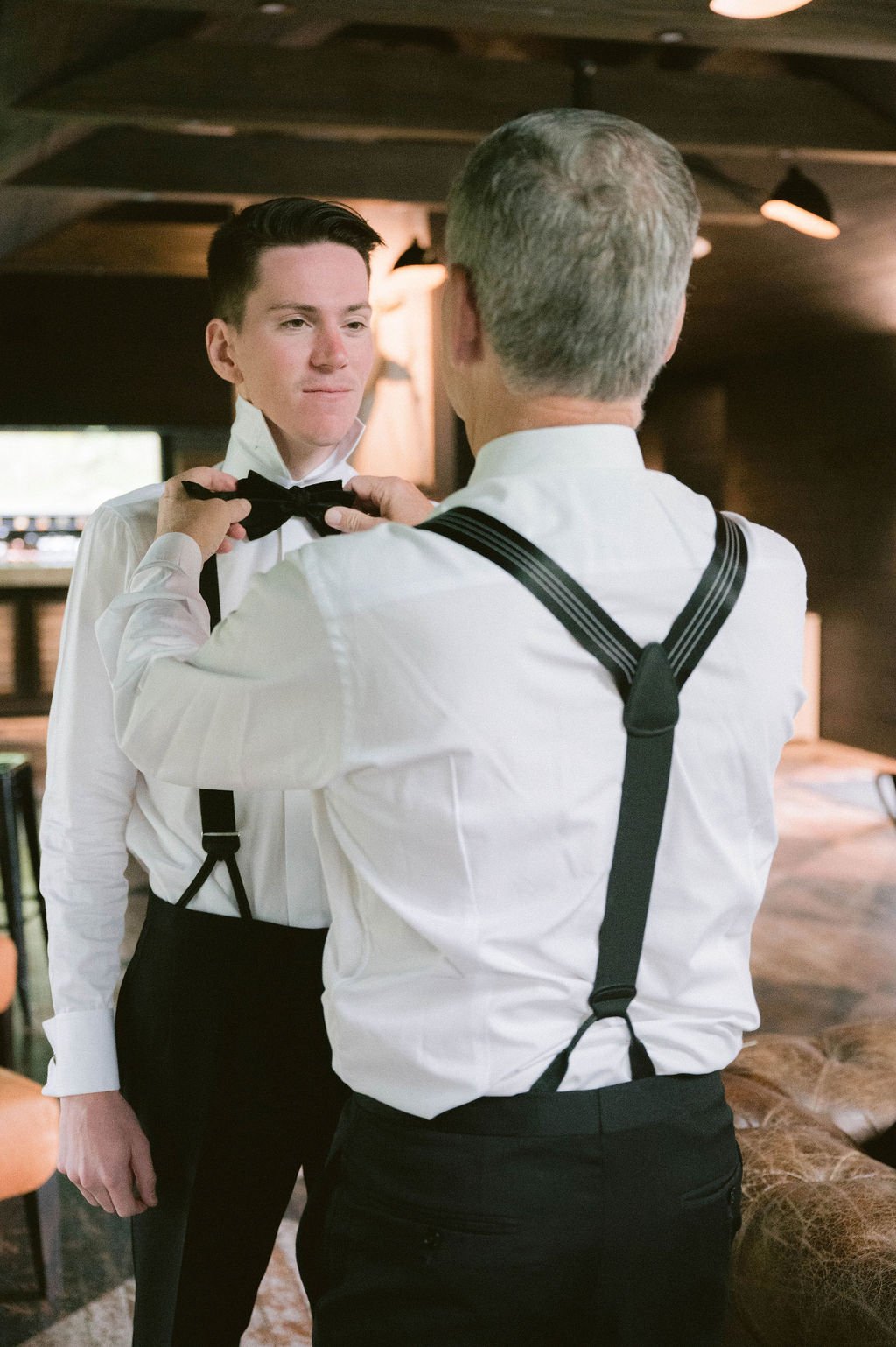 An older man helps a young man adjust his black bow tie. Both are dressed in formal attire, with white shirts and black suspenders, indoors with warm lighting.