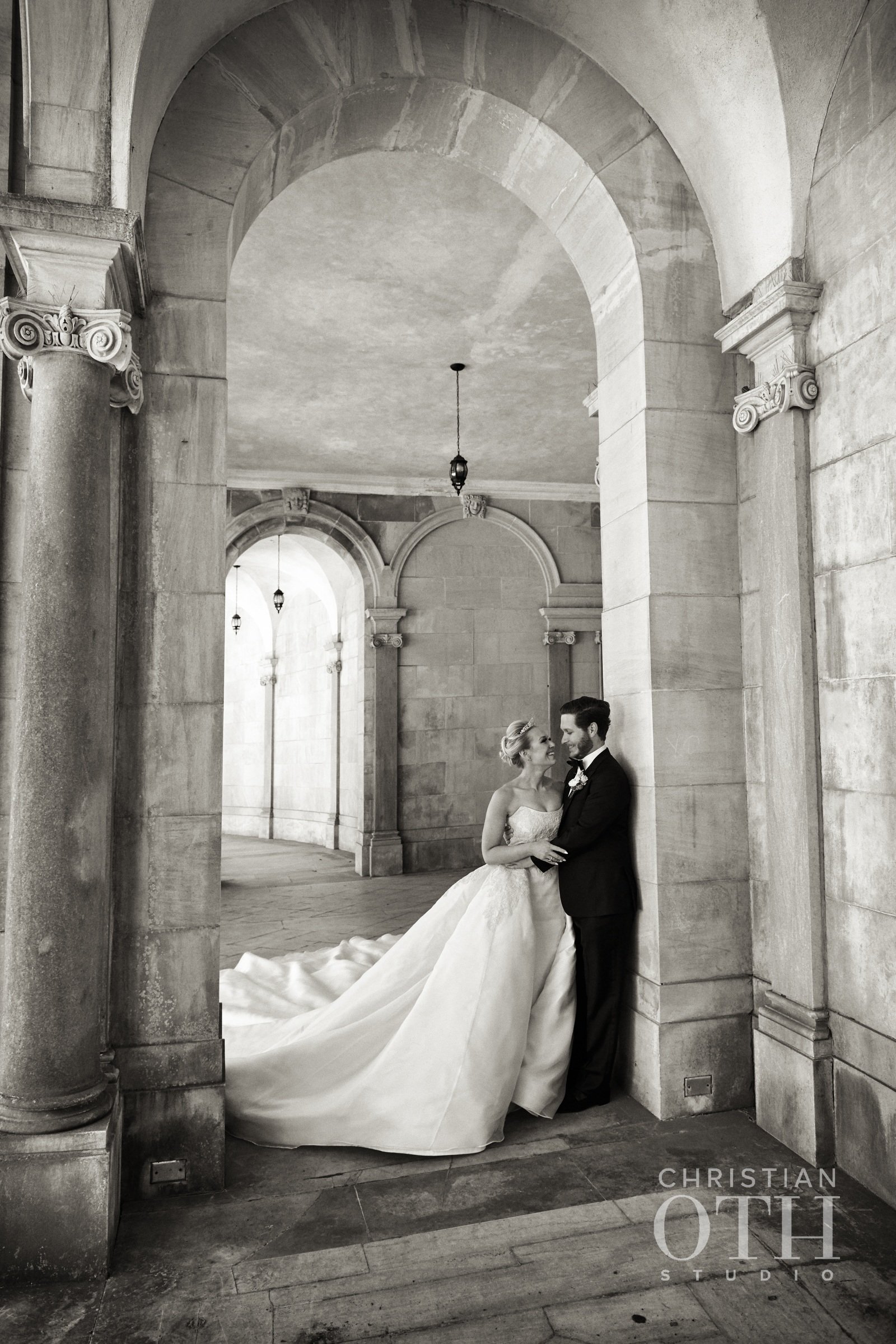 Bride and groom standing closely under an arched stone corridor, with the bride wearing a long wedding gown and the groom in a black suit, sharing a moment of affection.