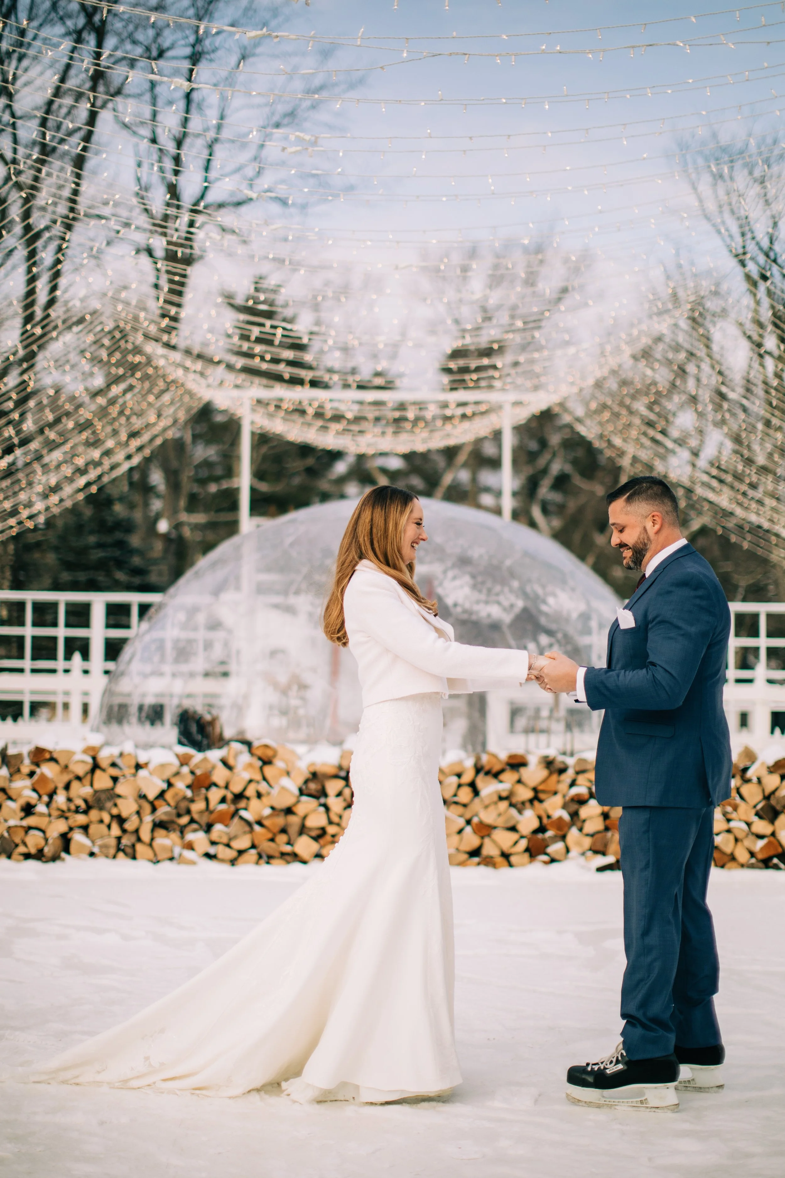 A bride and groom standing on ice, holding hands and smiling during a winter wedding ceremony outdoors. Behind them are stacked firewood, a clear dome, and string lights overhead.