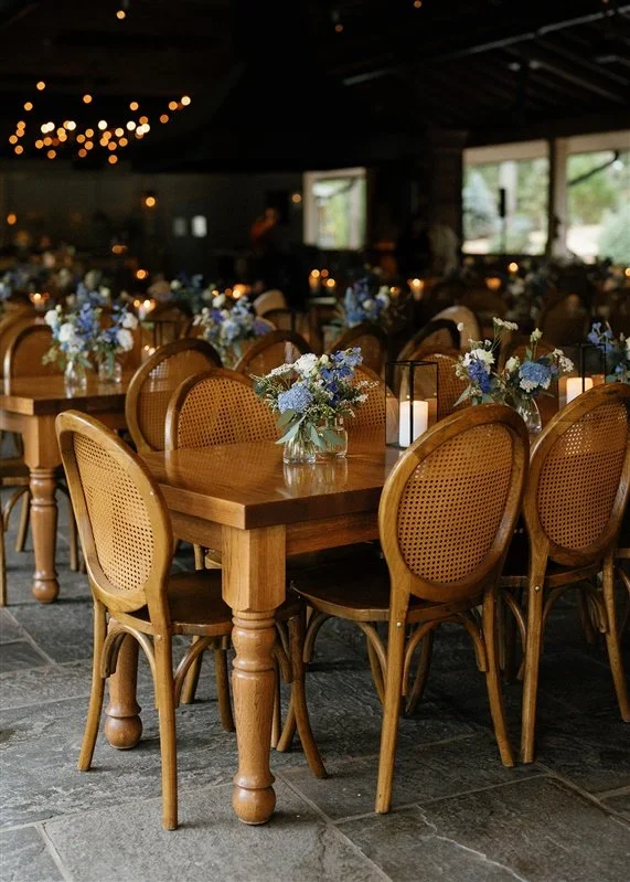 Indoor event space with wooden tables decorated with blue and white floral centerpieces and candles, set for a gathering or celebration.