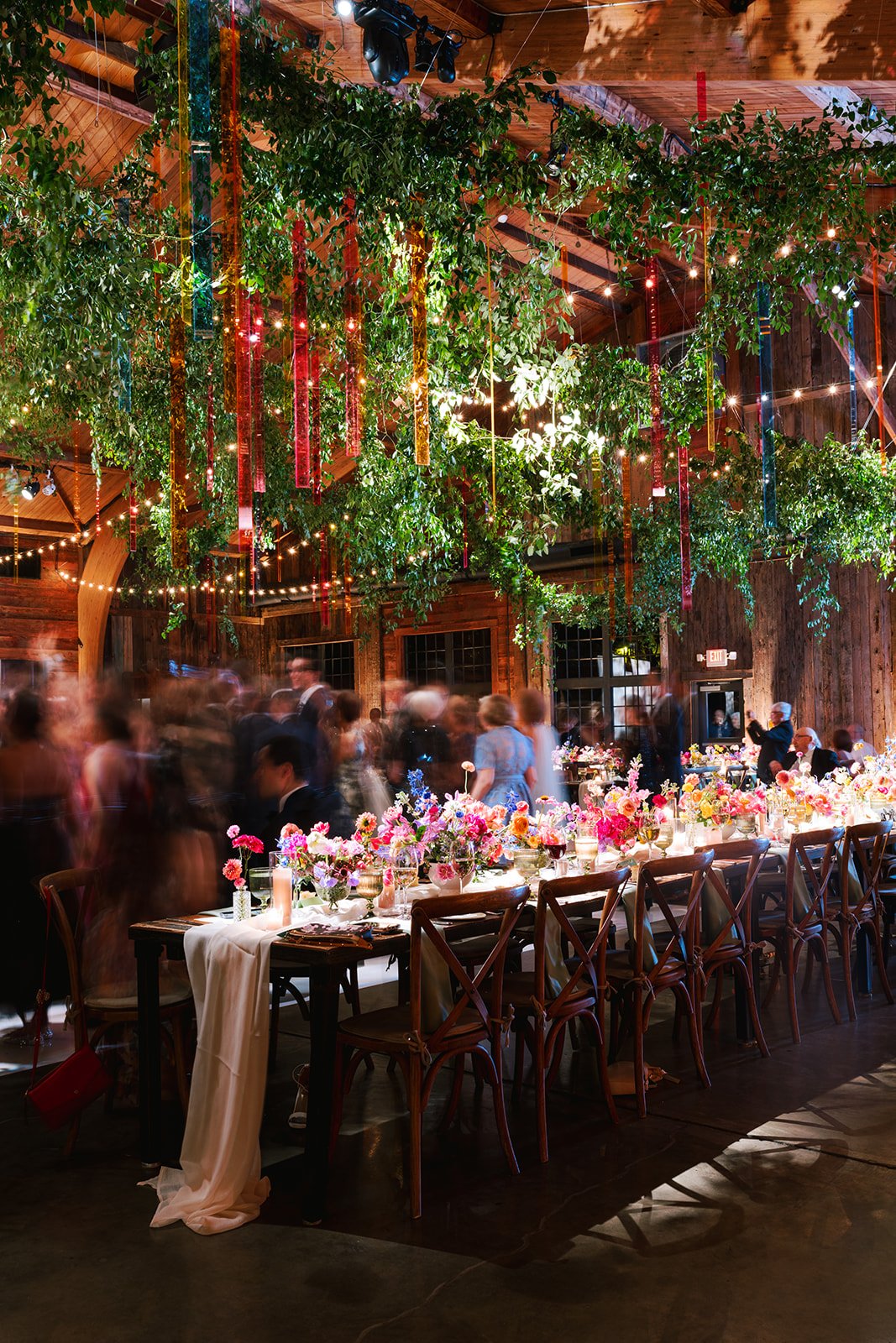 Inside a rustic wedding reception with wooden walls and ceiling, hanging greenery and colorful ribbons, decorated long table with floral centerpieces, candles, and place settings, surrounded by guests and ceiling string lights.