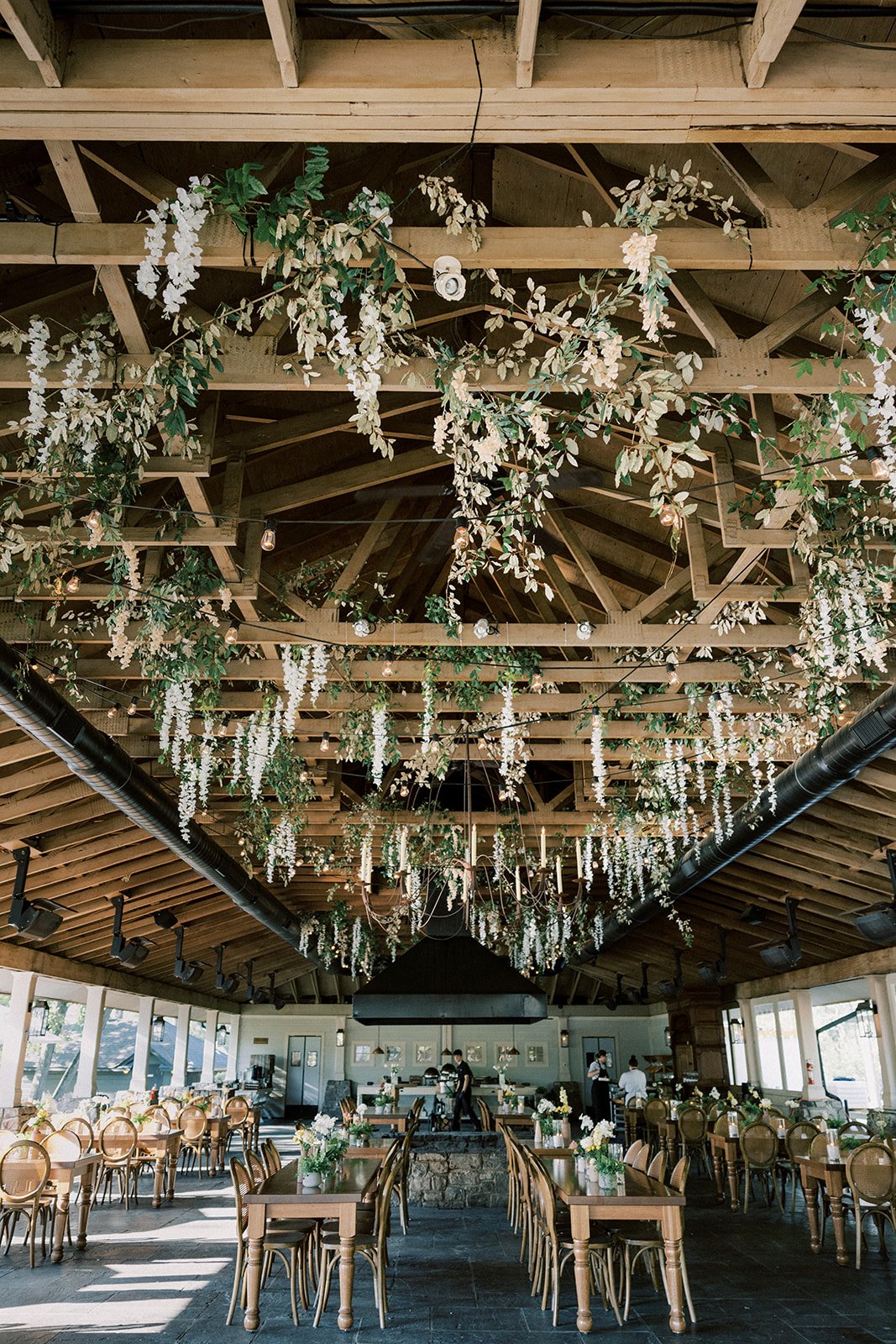 Interior of a decorated event space with wooden tables, chairs, hanging floral arrangements, string lights, and a black fireplace at the far end.