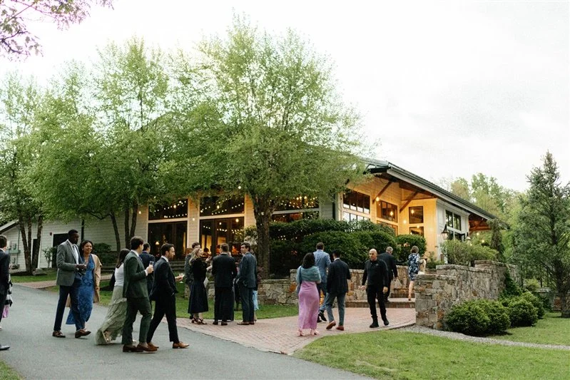 People dressed in formal attire gathering outside a house with string lights and a stone pathway in a lush, green yard during evening.