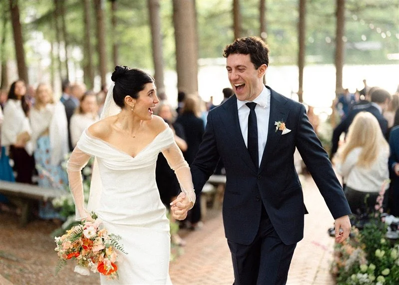A bride and groom walking hand-in-hand and smiling at each other at their wedding ceremony outdoors in a wooded area with guests in the background.