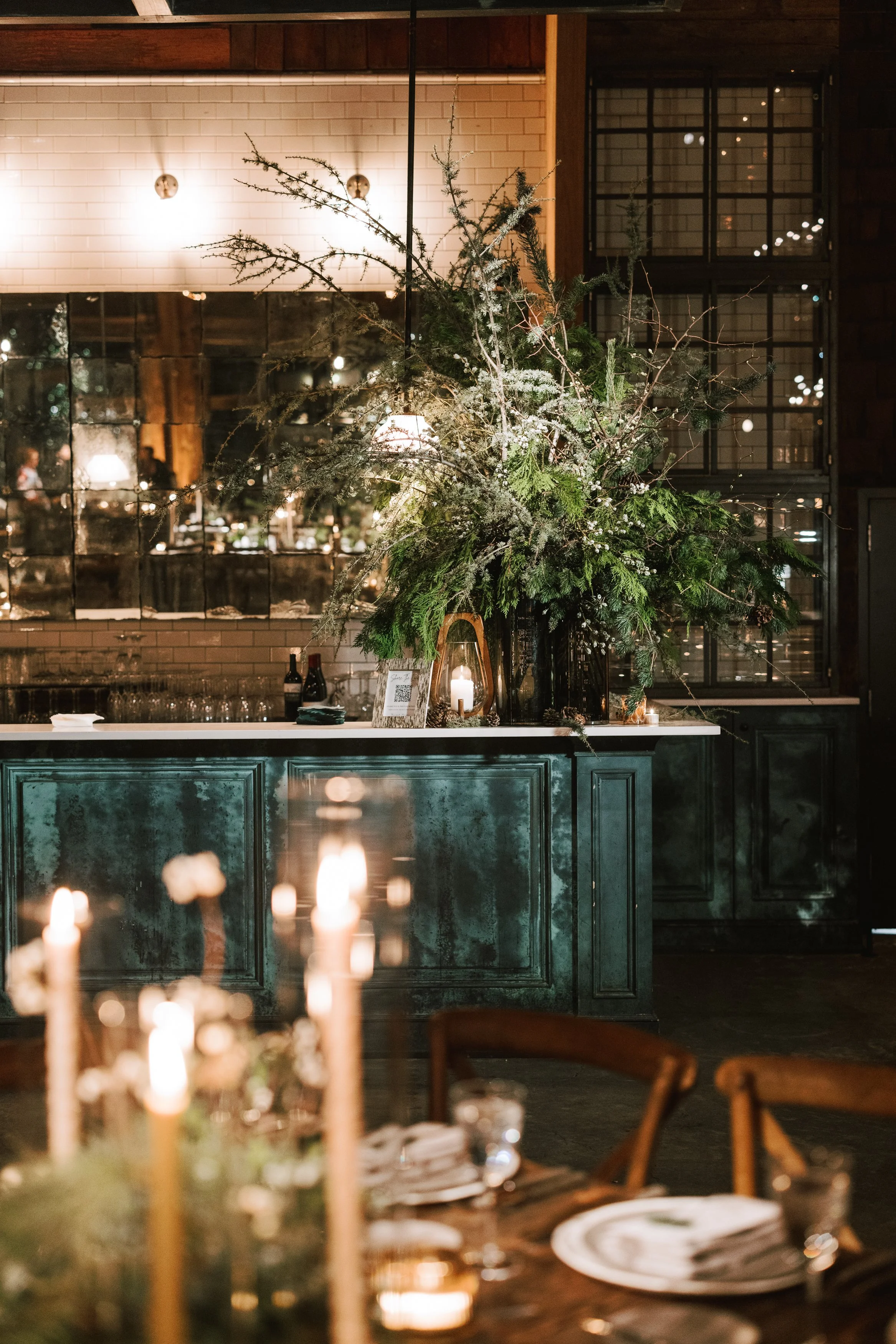 Elegant indoor dining setting with a dark green sideboard topped with a large, lush Christmas greenery arrangement, including pine branches, snow-dusted twigs, and white flowers. Decorative lantern with a candle and small wine bottles are on the side