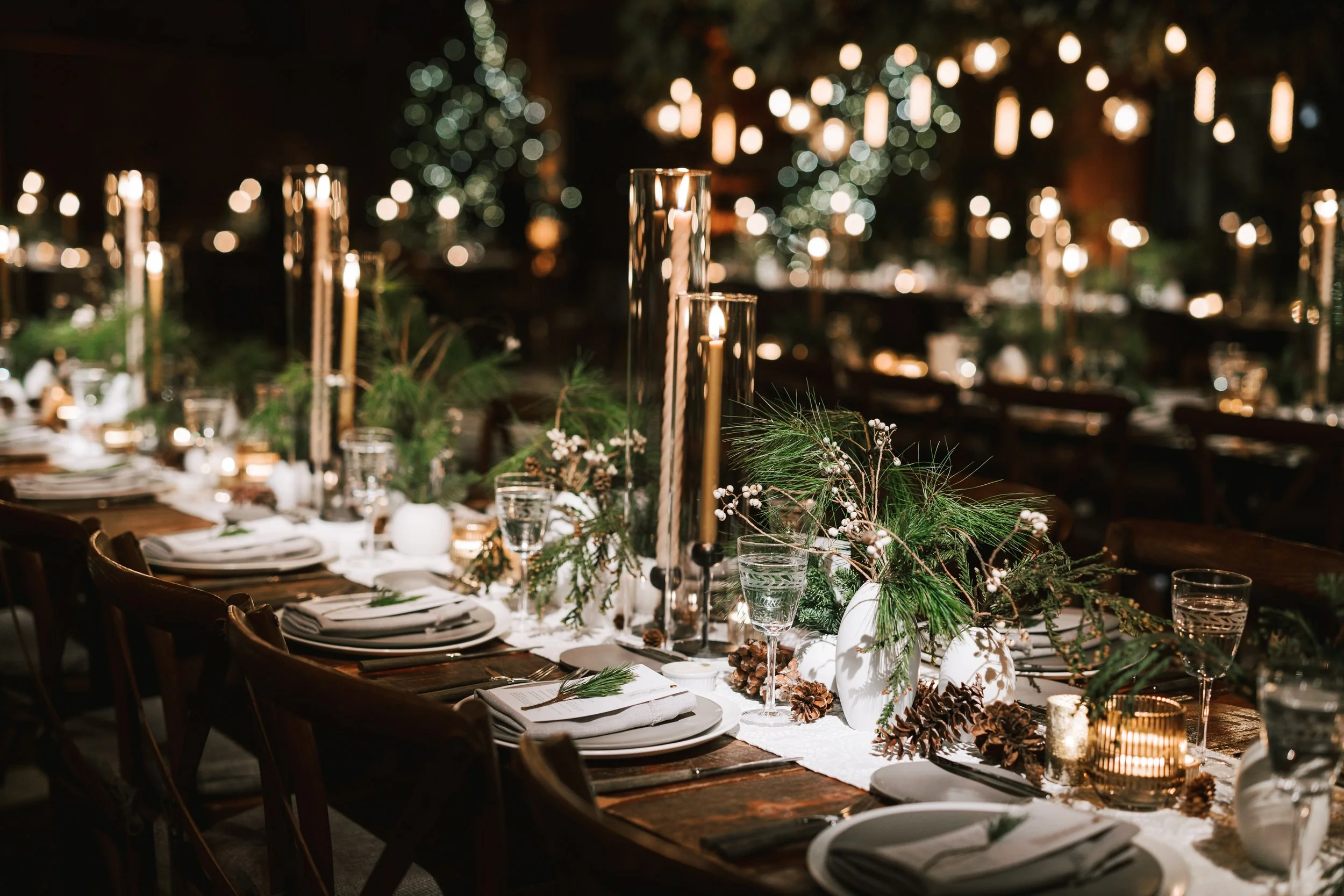 A beautifully decorated Christmas dinner table with greenery, pinecones, white vases, candles, and glasses of water, set in a warmly lit, festive atmosphere.