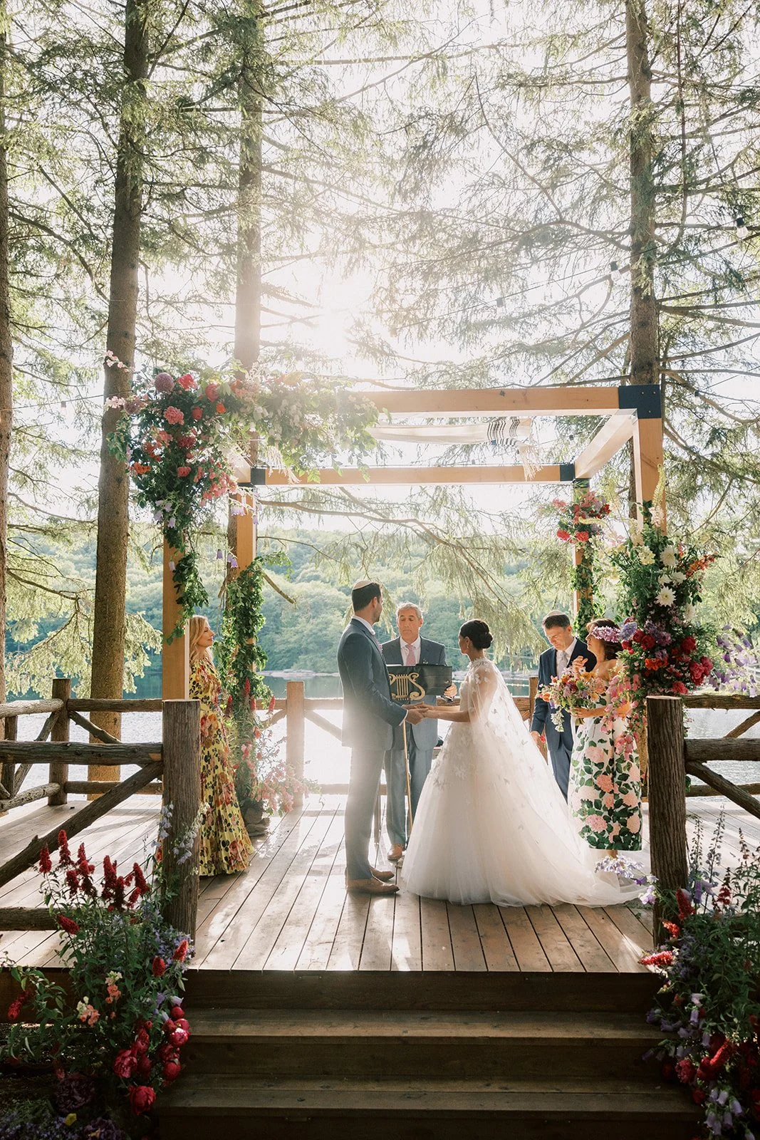 A wedding ceremony taking place outdoors on a wooden platform surrounded by trees and water, with the bride and groom exchanging vows under a floral arch.