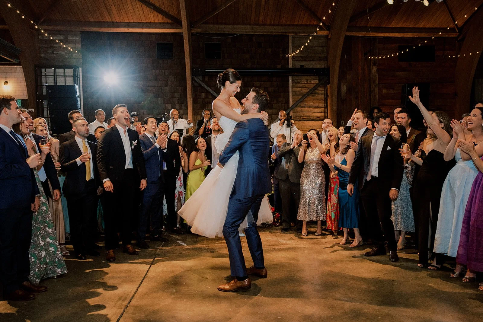 A wedding reception with a bride and groom dancing, surrounded by friends and family in a rustic indoor venue with wooden beams and string lights.