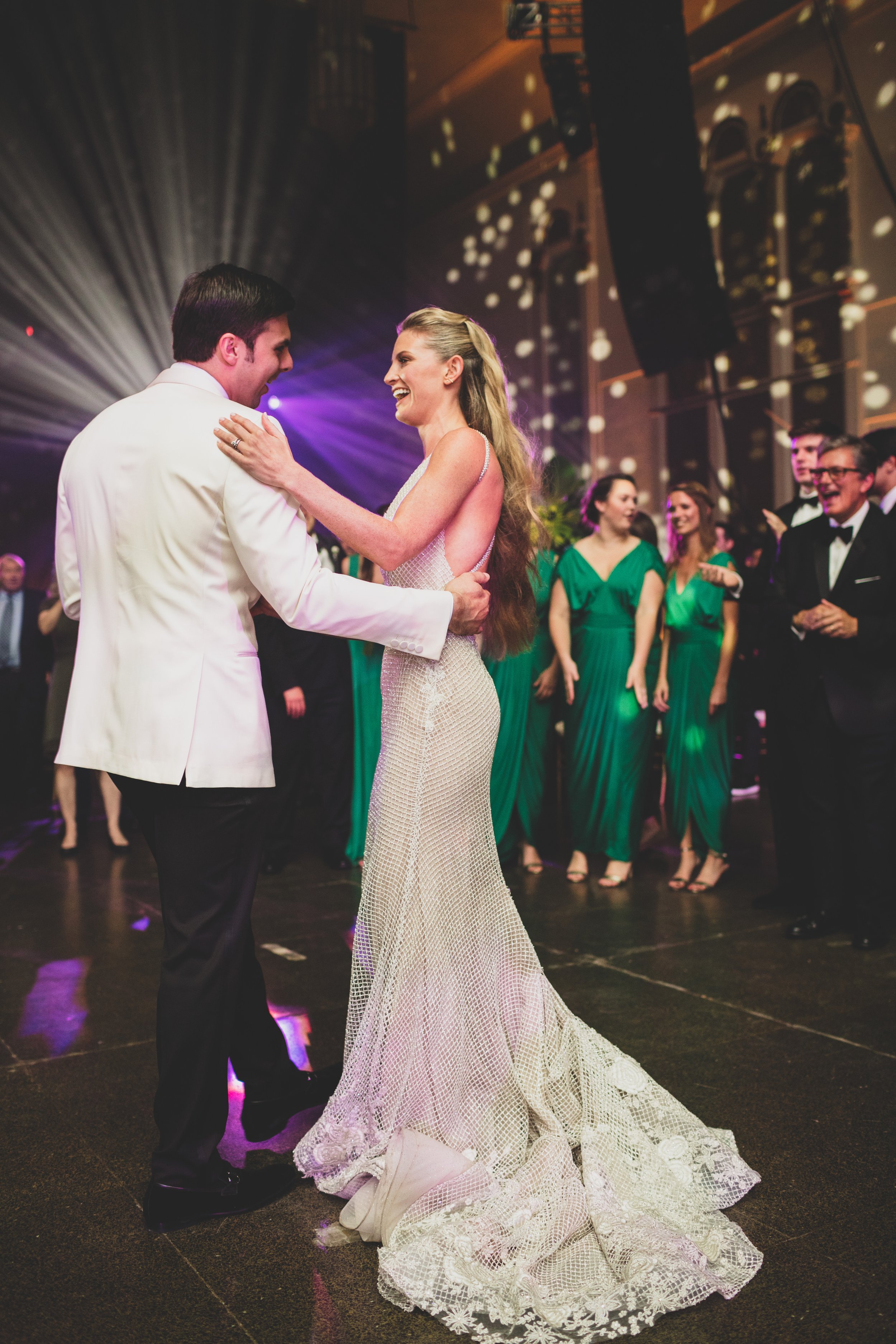 A bride and groom dancing at their wedding reception, surrounded by guests in formal attire and green dresses, with colorful lighting and decorations.