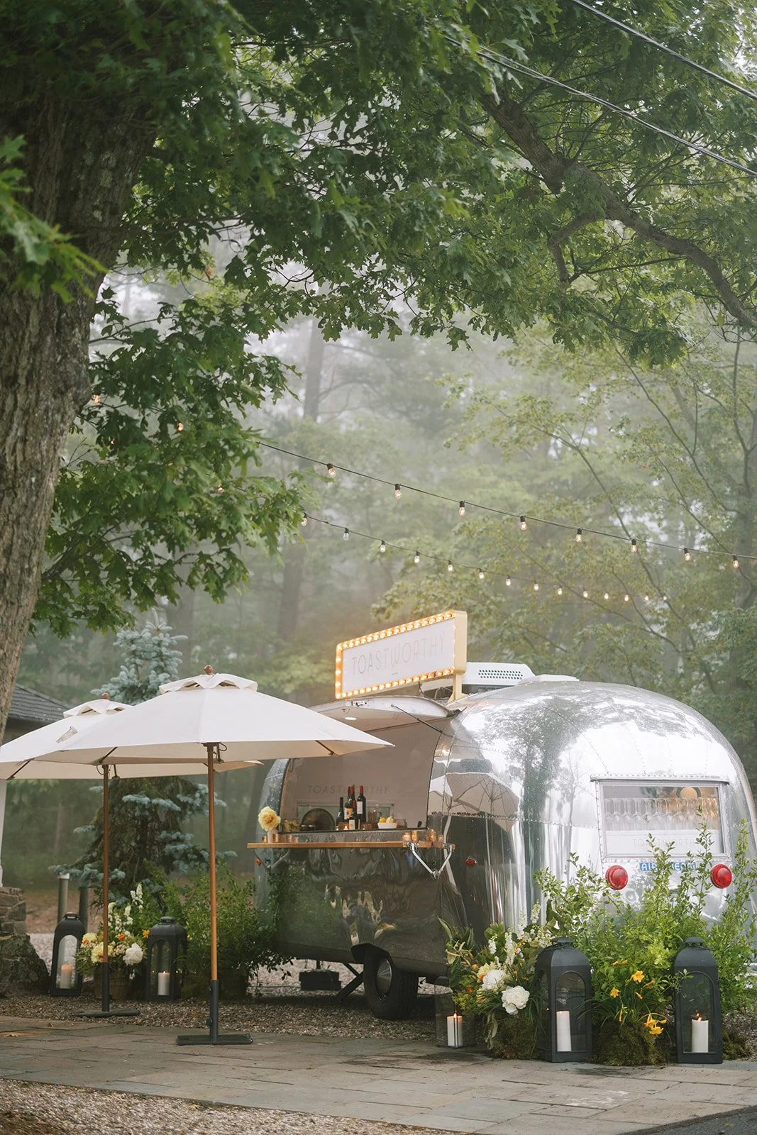 A shiny silver camper trailer decorated with flowers, candles, and lanterns, set up outdoors under green trees with string lights overhead. There is a white umbrella and a sign that says 'TOASTWORTHY' on top of the trailer, indicating it is likely a 