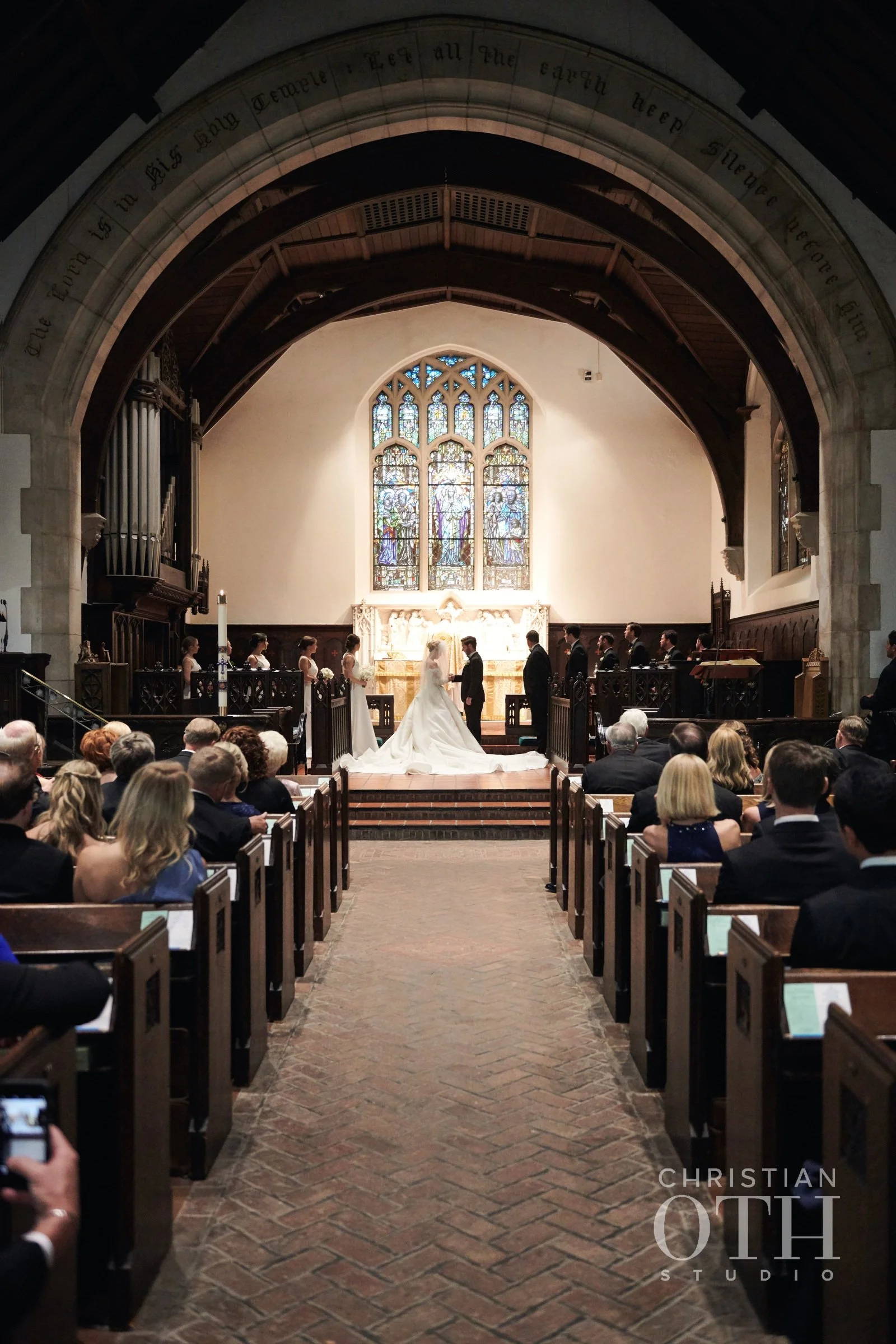 A wedding ceremony takes place inside a church with a bride and groom standing at the altar, surrounded by bridesmaids and groomsmen. The church features a stained glass window behind the altar, wooden pews filled with seated guests, and an arched ce