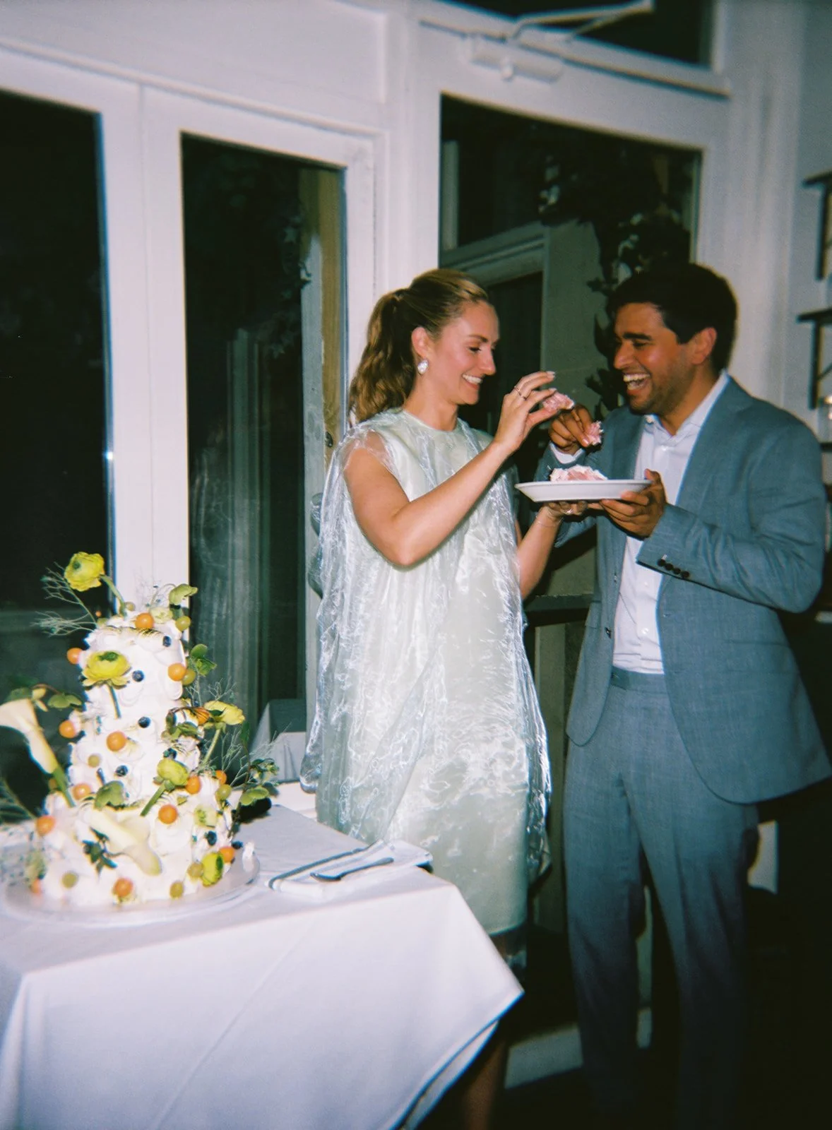 A wedding reception with a woman in a white dress and a man in a gray suit sharing cake, smiling, near a decorated cake on a table.