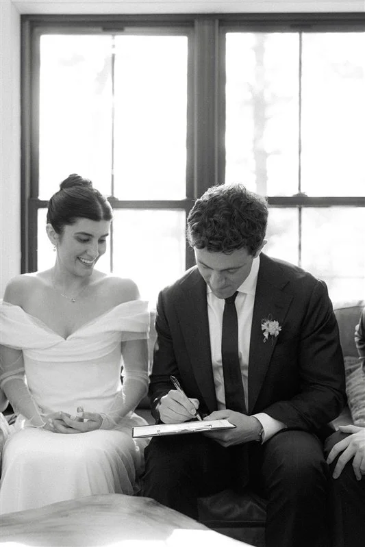 A bride and groom at their wedding ceremony, with the groom signing a marriage document.