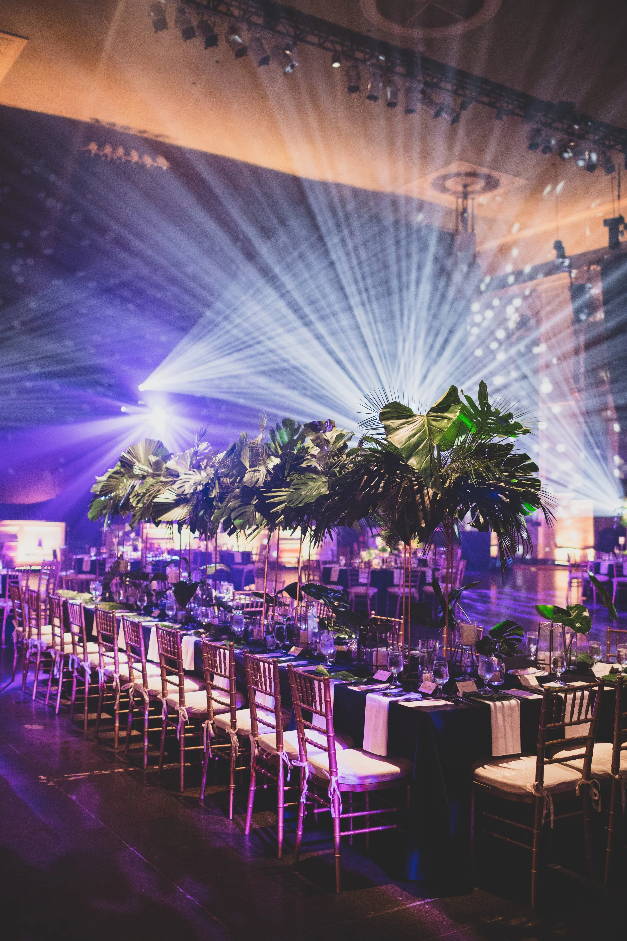 Elegant event setup with a long dining table decorated with lush greenery and gold chairs, illuminated by stage lighting with purple and white beams, in a large indoor venue.
