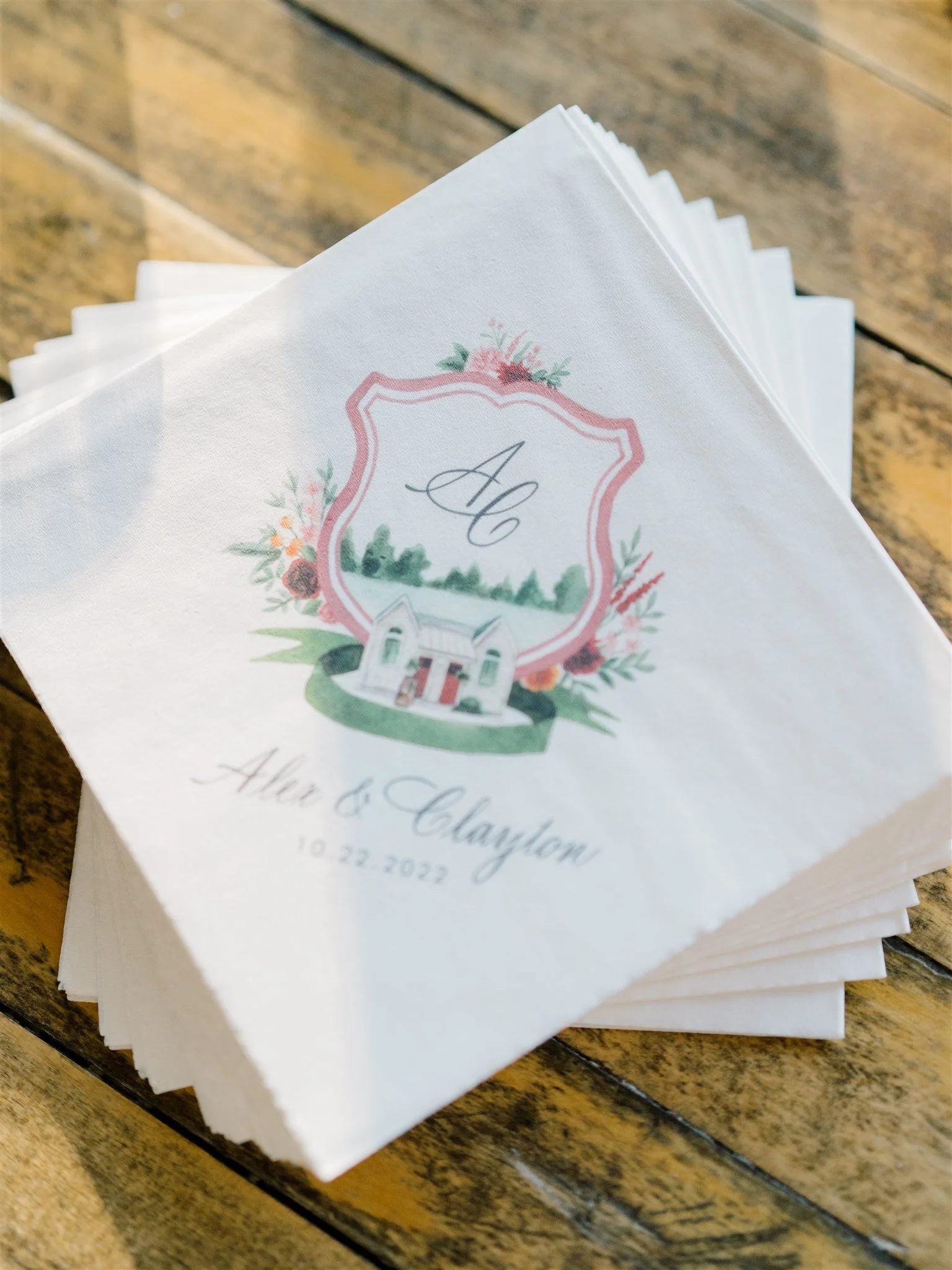 Wedding napkins with floral design, monogram, and the names Alice & Clayton, dated 10-22-2022, placed on a wooden surface.