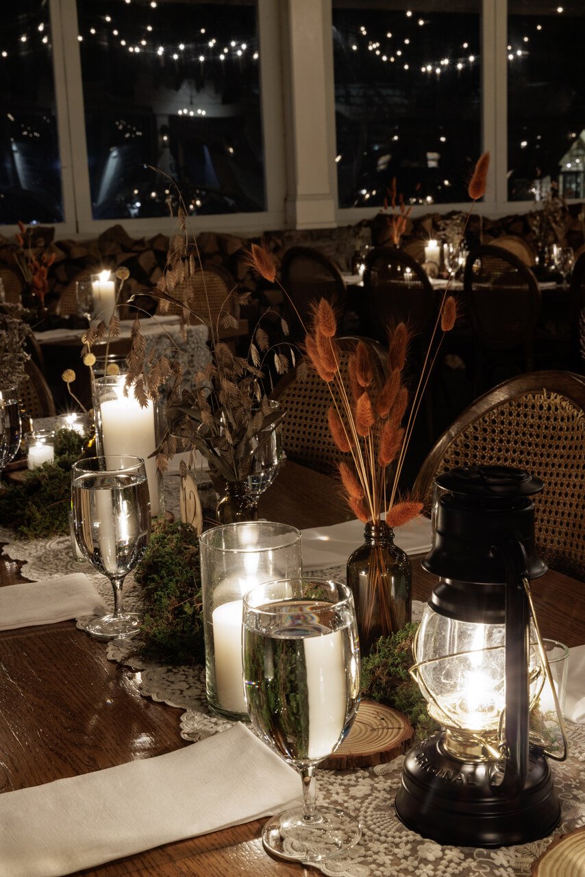Elegant dinner table with candles, glassware, dried flowers, and lanterns in a warmly lit setting.