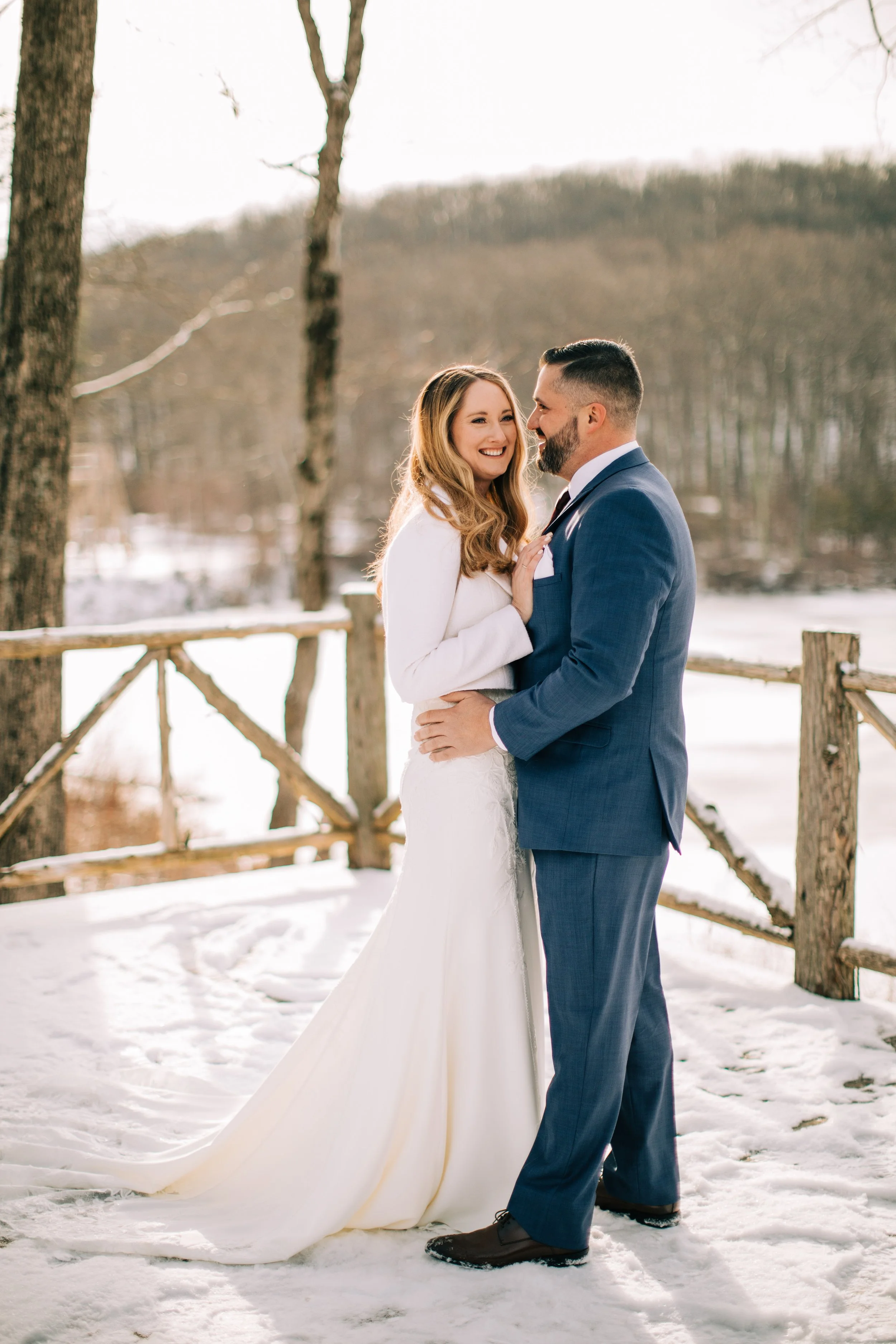 A bride and groom in wedding attire standing on snow-covered ground outdoors, smiling and embracing each other with bare trees and a wooden fence in the background.