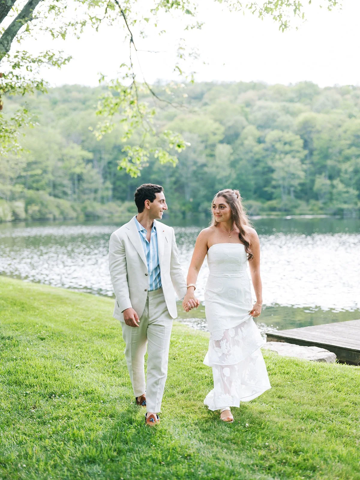 A woman in a white strapless wedding dress and a man in a white suit walking hand in hand along a grassy area by a lake with trees in the background.