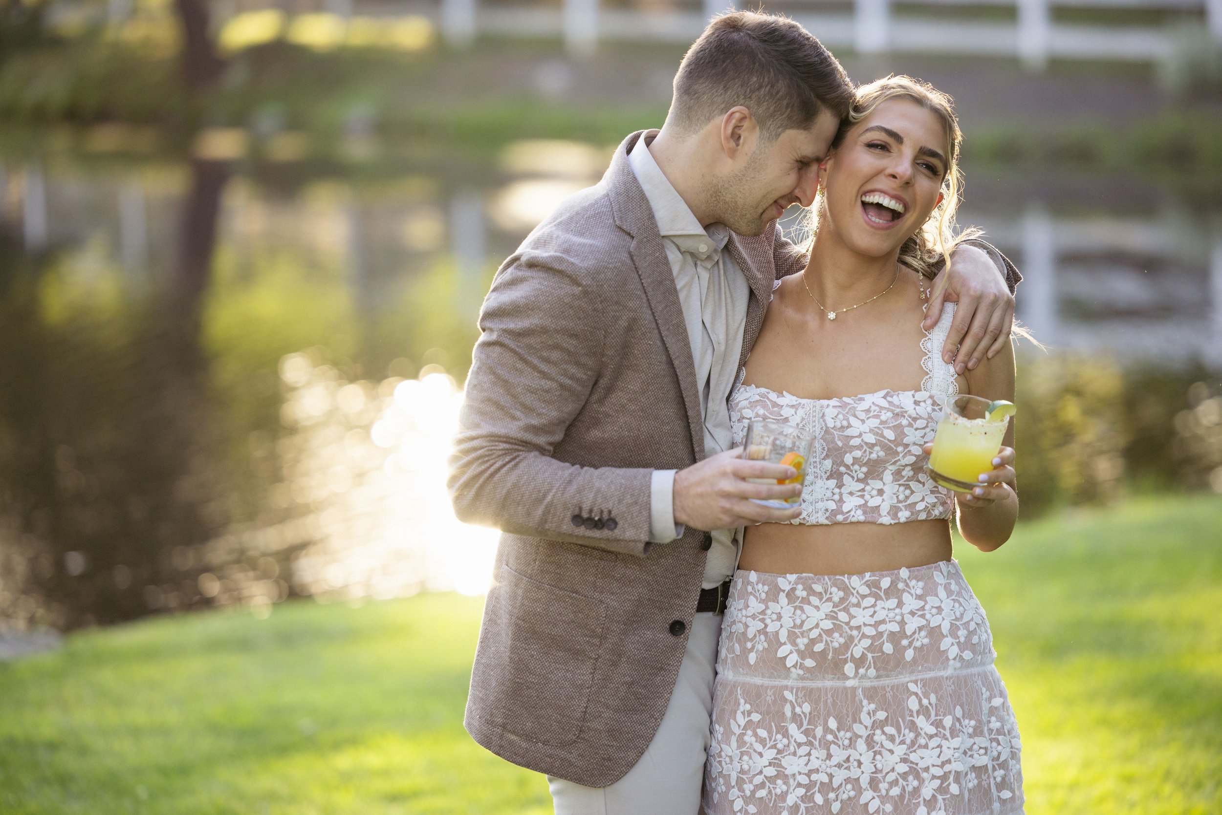 A couple near a lake, smiling and holding drinks, enjoying a warm outdoor setting.