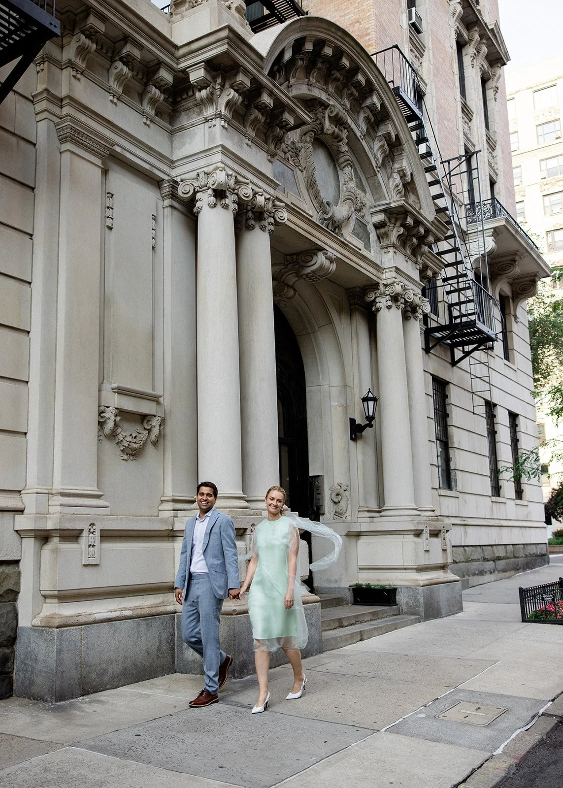 A smiling couple walking hand in hand in front of a historic building with ornate stone architecture, large columns, and fire escape ladders, on a city sidewalk.