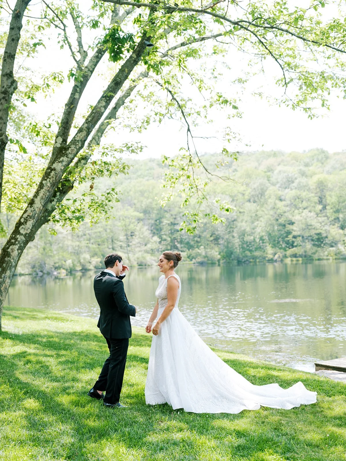 A bride and groom standing by a lake, with lush green trees and hills in the background, under a large tree with bright green leaves, on a sunny day.