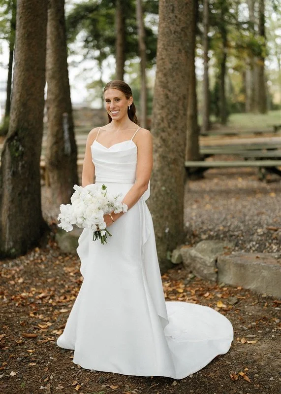 A bride in a white wedding dress holding a bouquet of white flowers, standing outdoors among trees.