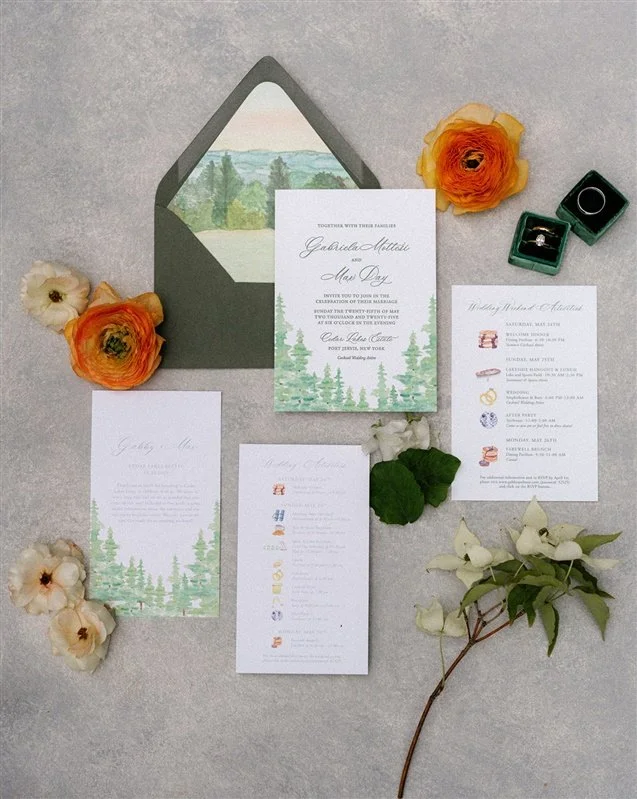 Wedding invitation suite with floral accents, including flowers, greenery, and rings, arranged on a light gray surface.