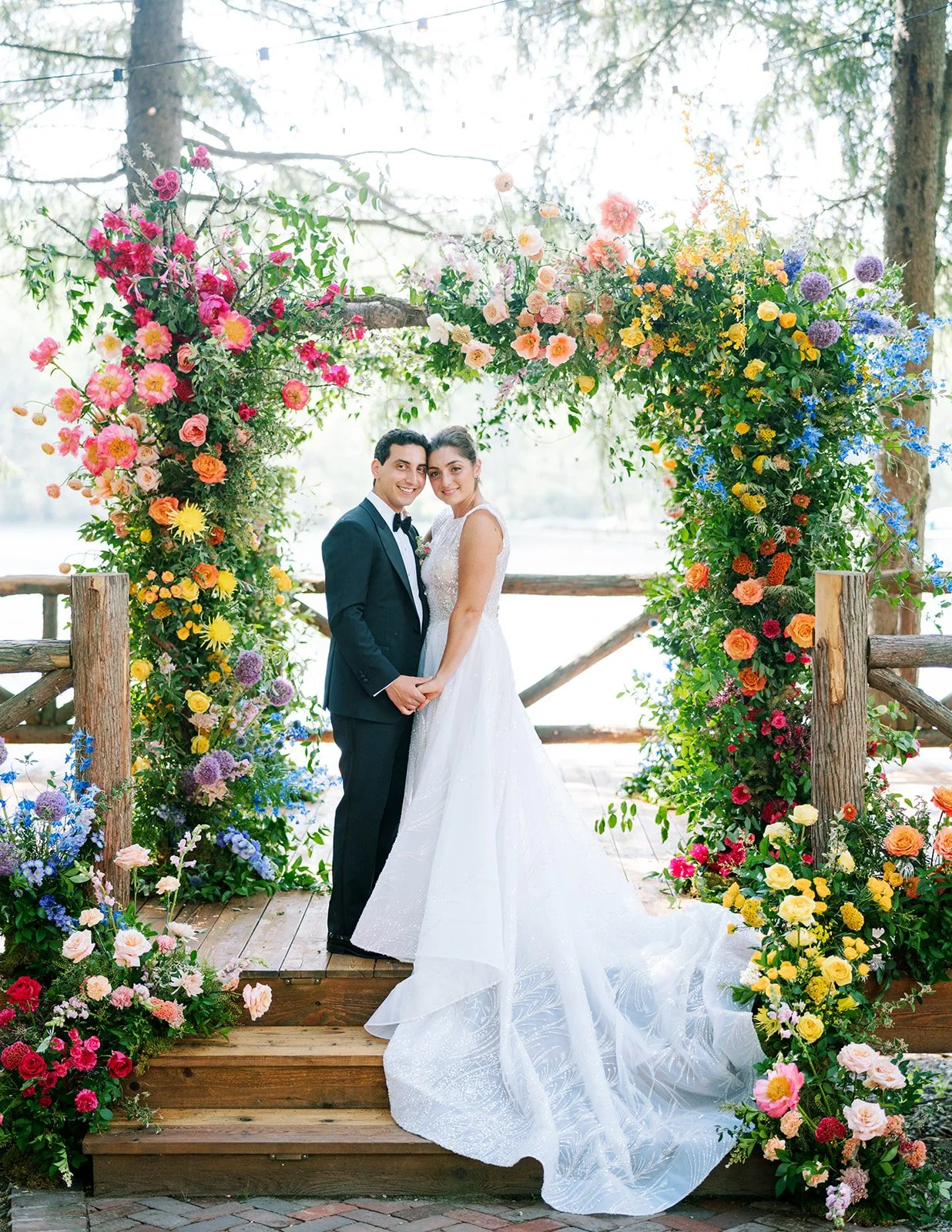 A bride and groom standing under a floral arch on a wooden platform.
