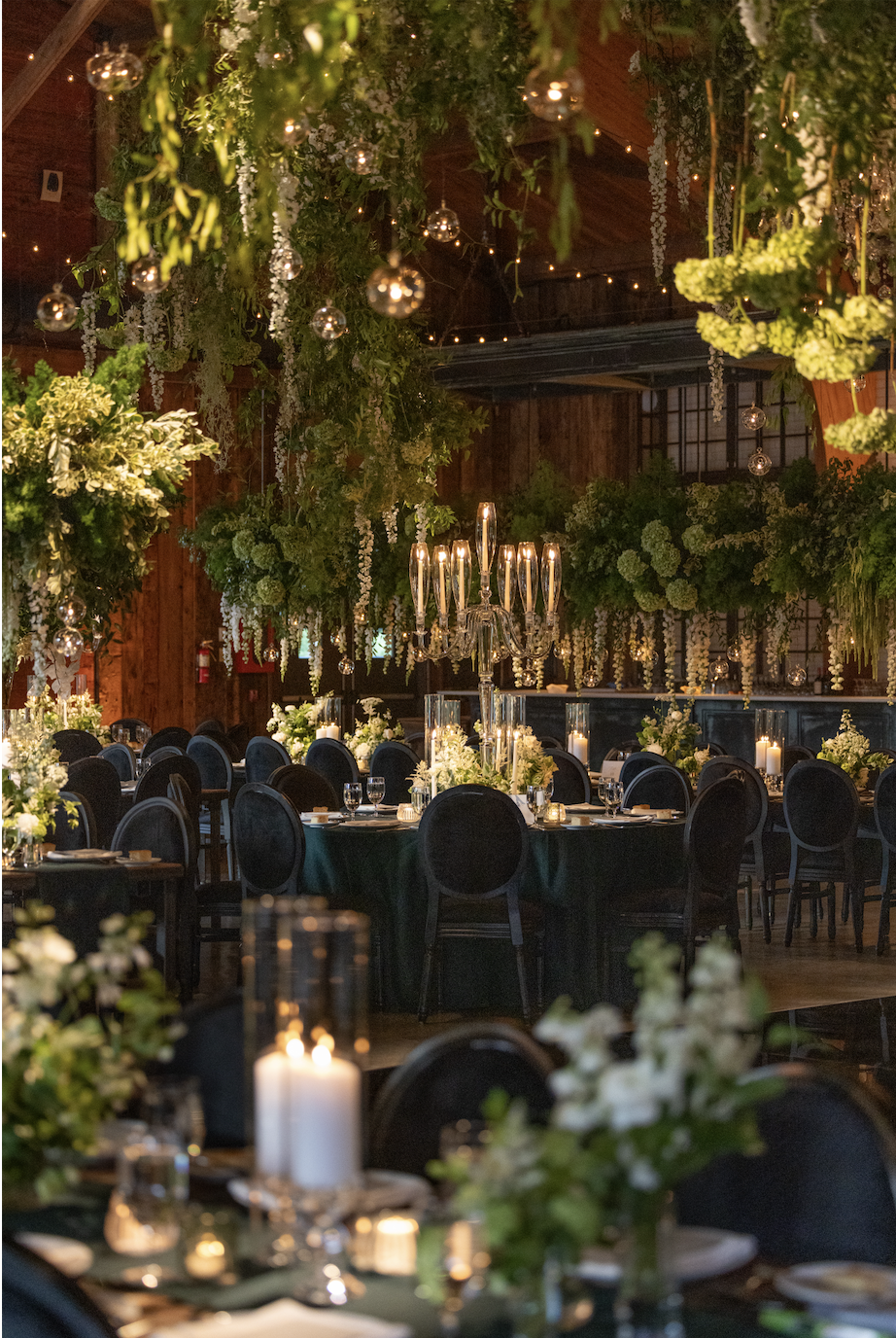 Elegant indoor wedding reception with round tables, black chairs, lush green and white floral centerpieces, candles, and hanging greenery and glass ornaments, under a wooden ceiling with warm lighting.
