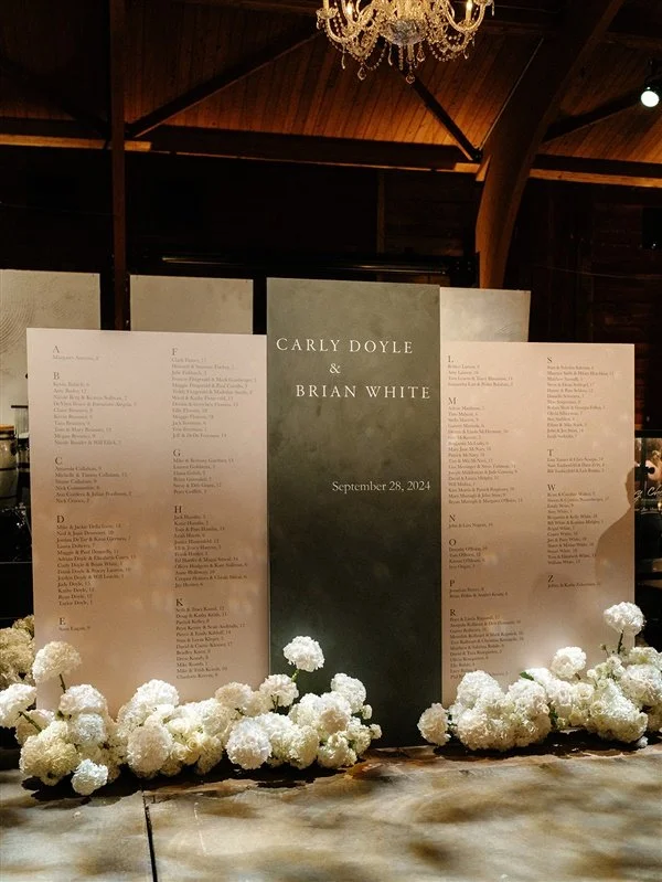 Wedding reception seating chart with a black center panel displaying the names Carly Doyle and Brian White, and the date September 28, 2024. The chart is flanked by flowers and is set inside a wooden barn with a chandelier overhead.