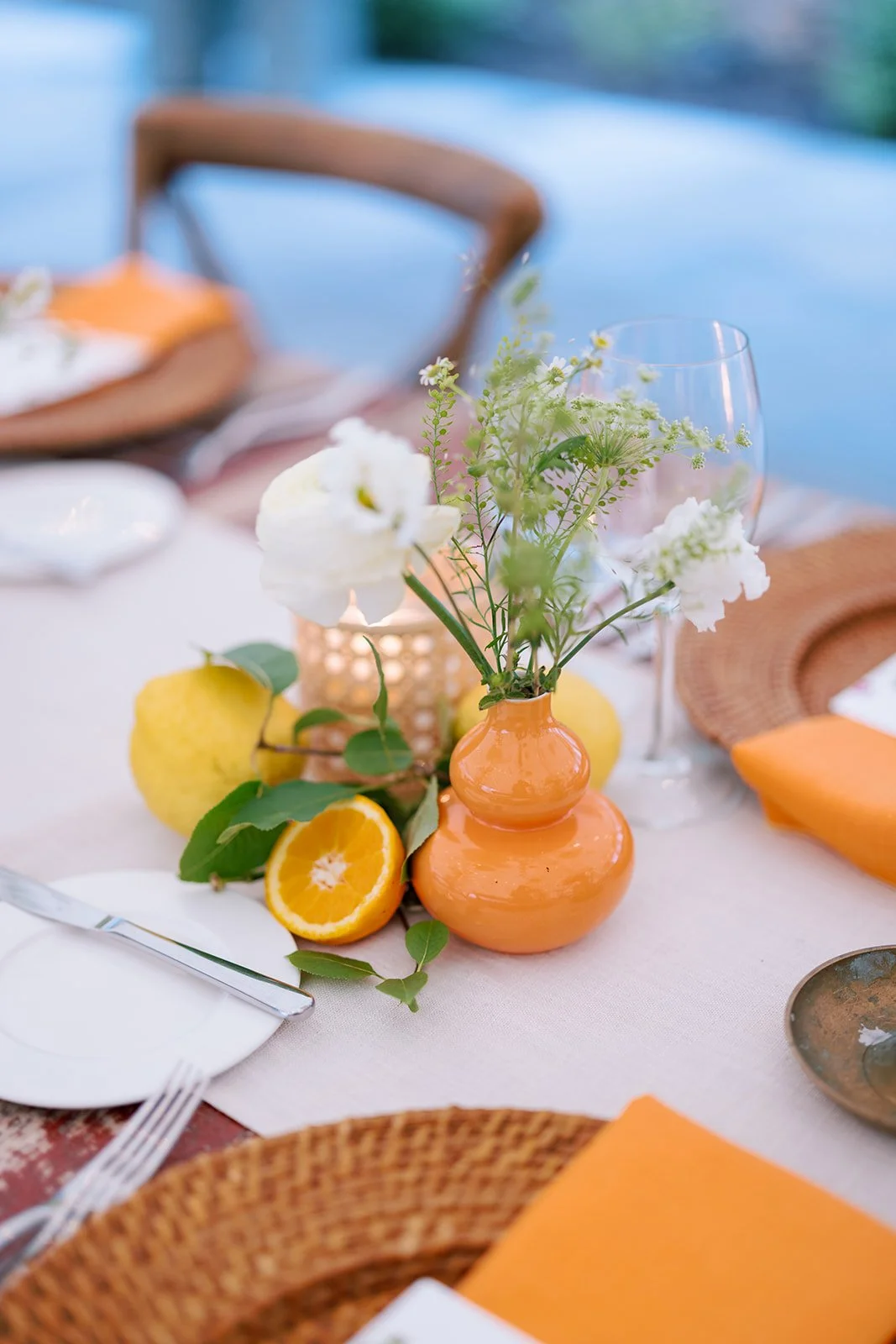 A table setting with orange-themed decorations, including a small orange vase with white flowers, citrus fruits like sliced orange and lemon, and orange napkins, on a white tablecloth with place settings and wicker placemats.
