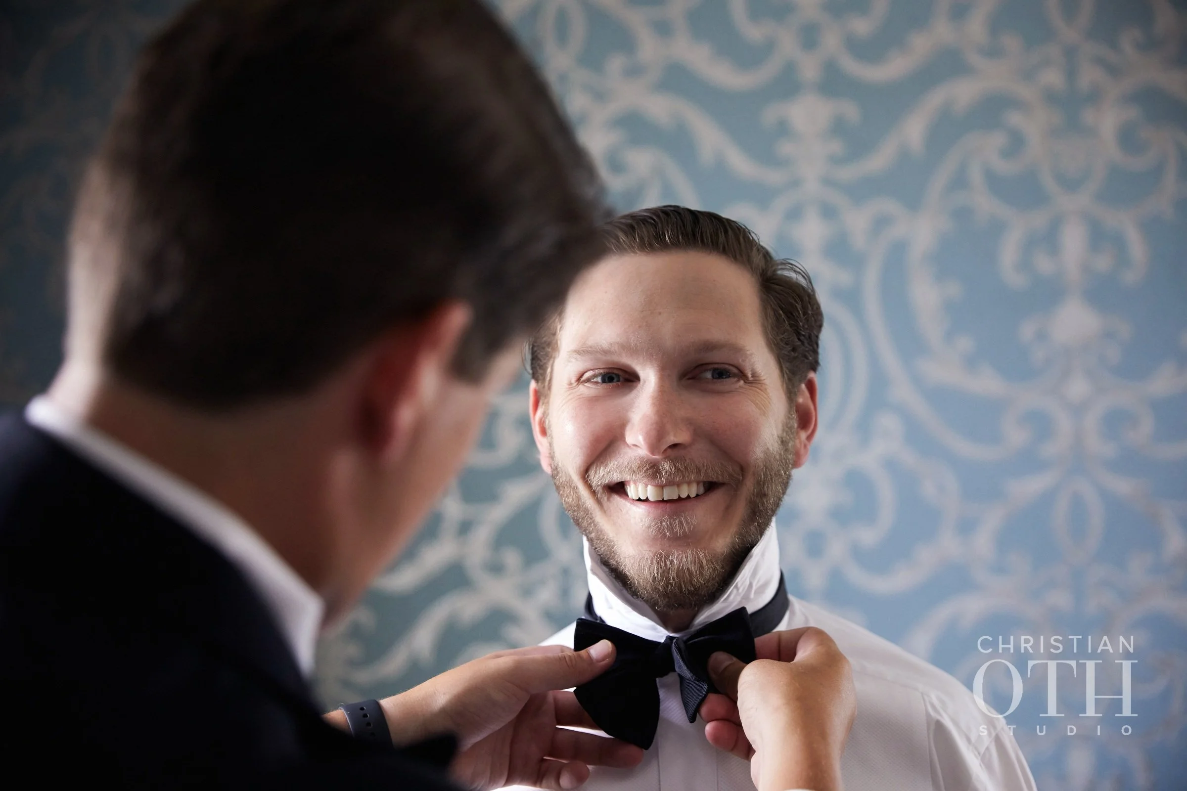 A man in formal attire being helped with his black bow tie, smiling, in front of a patterned blue and white wallpaper.