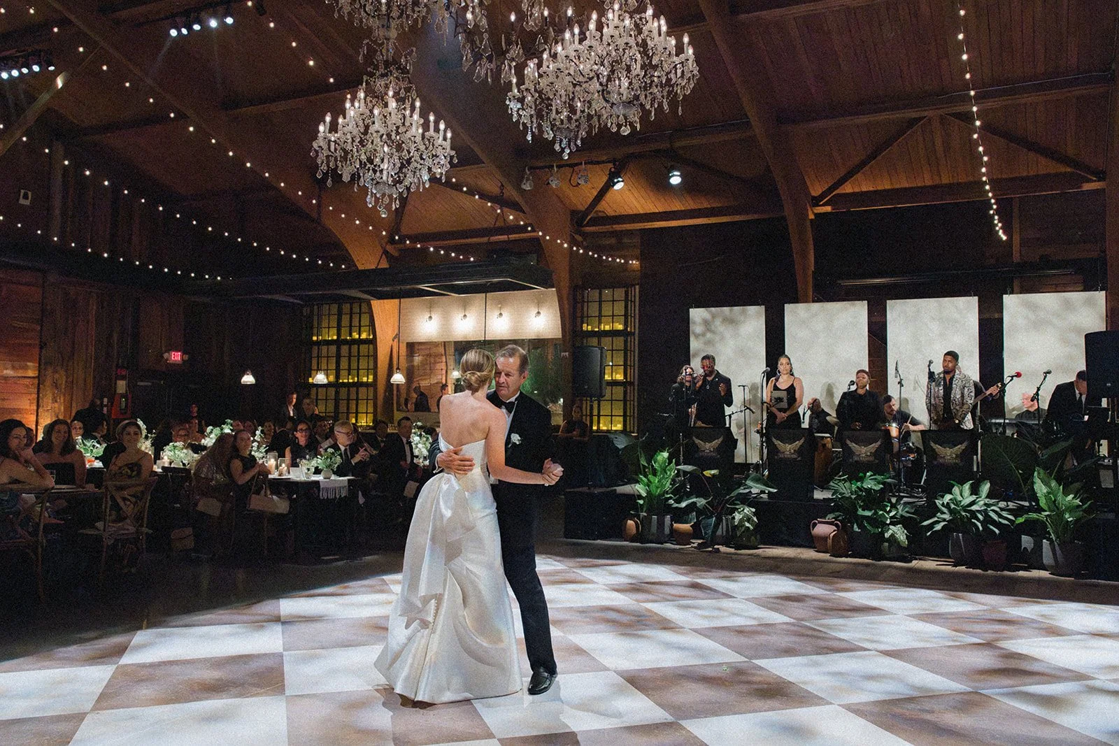 Bride and groom dancing at their wedding reception, with guests seated at tables and a band performing on stage in a decorated indoor venue with chandeliers and string lights.