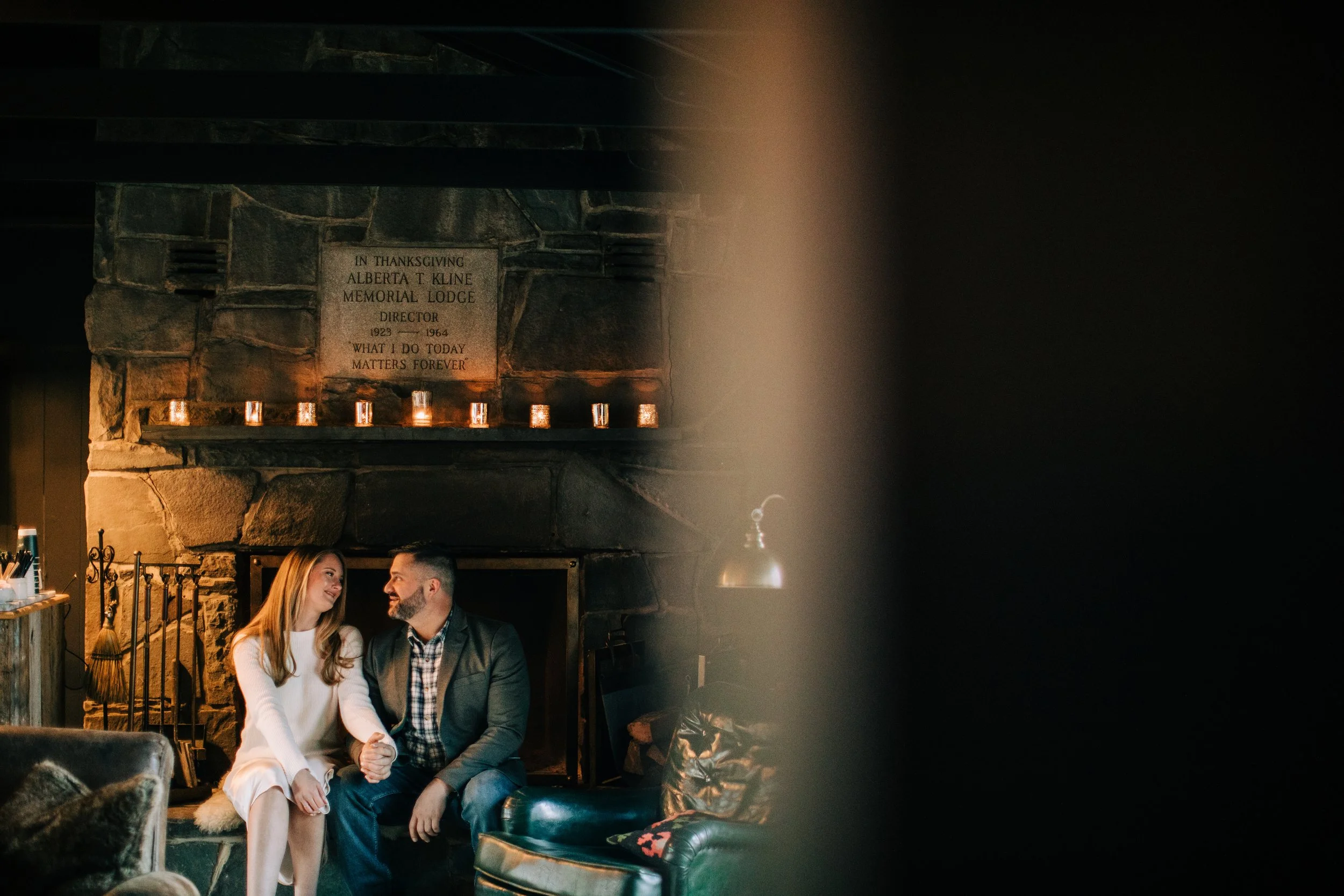 A couple sitting close together on a couch, smiling and holding hands in a cozy, dimly lit room with a stone fireplace and a memorial plaque on the wall.