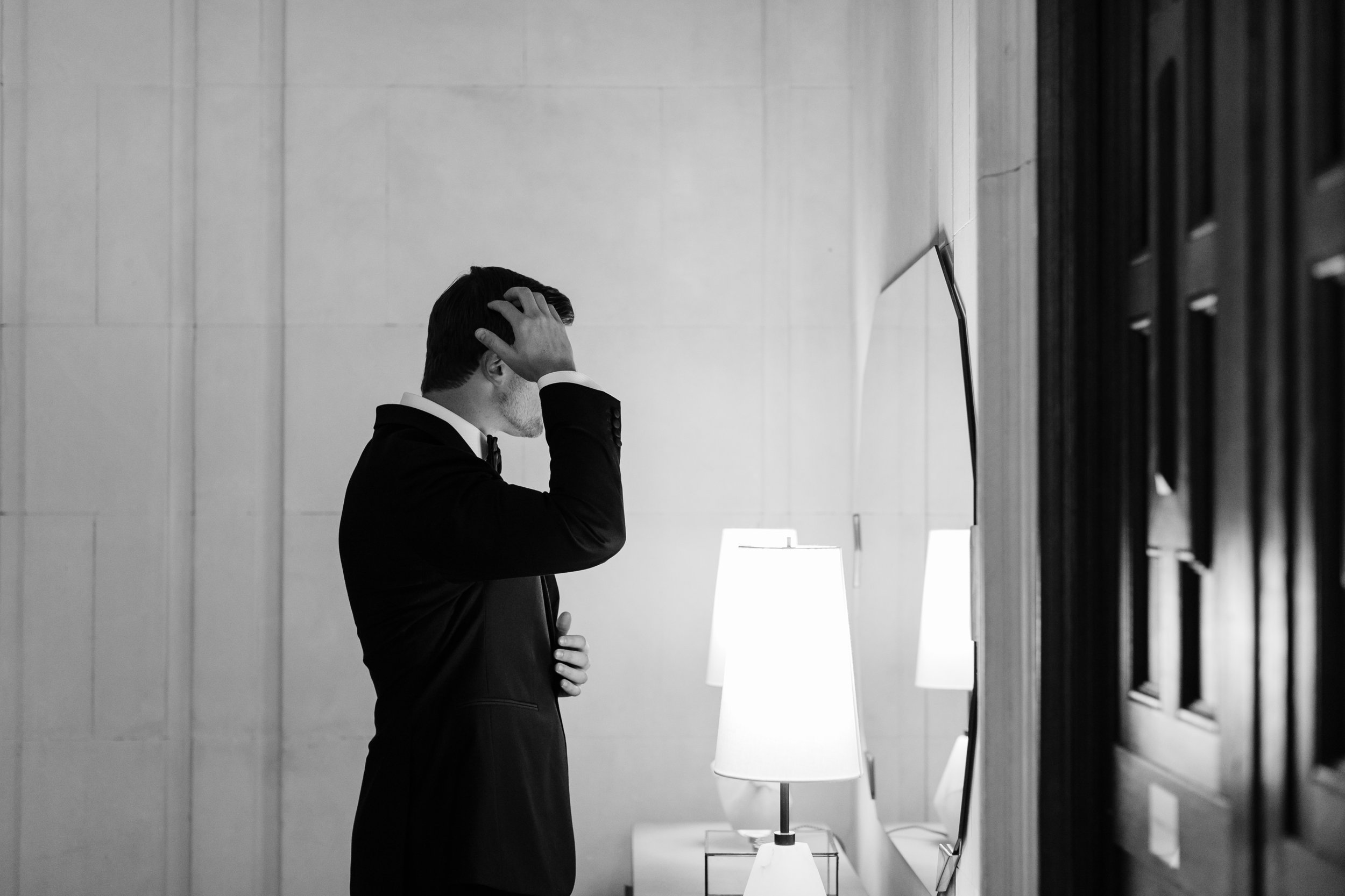 A man in a tuxedo adjusting his hair while looking at a mirror in a modern, minimalist room with lamps and wall decor.