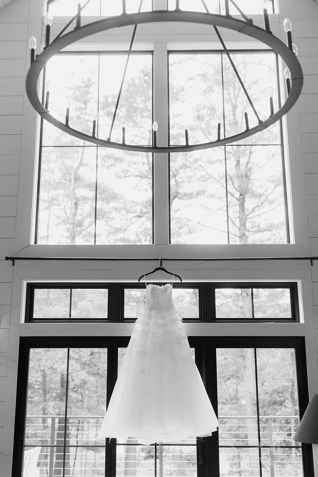 A wedding dress hanging in front of large windows in a bright room with a chandelier above.