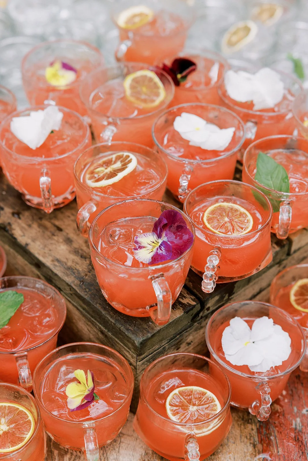 Multiple glasses of pink citrus drink garnished with lemon slices and edible flowers on a wooden tray.