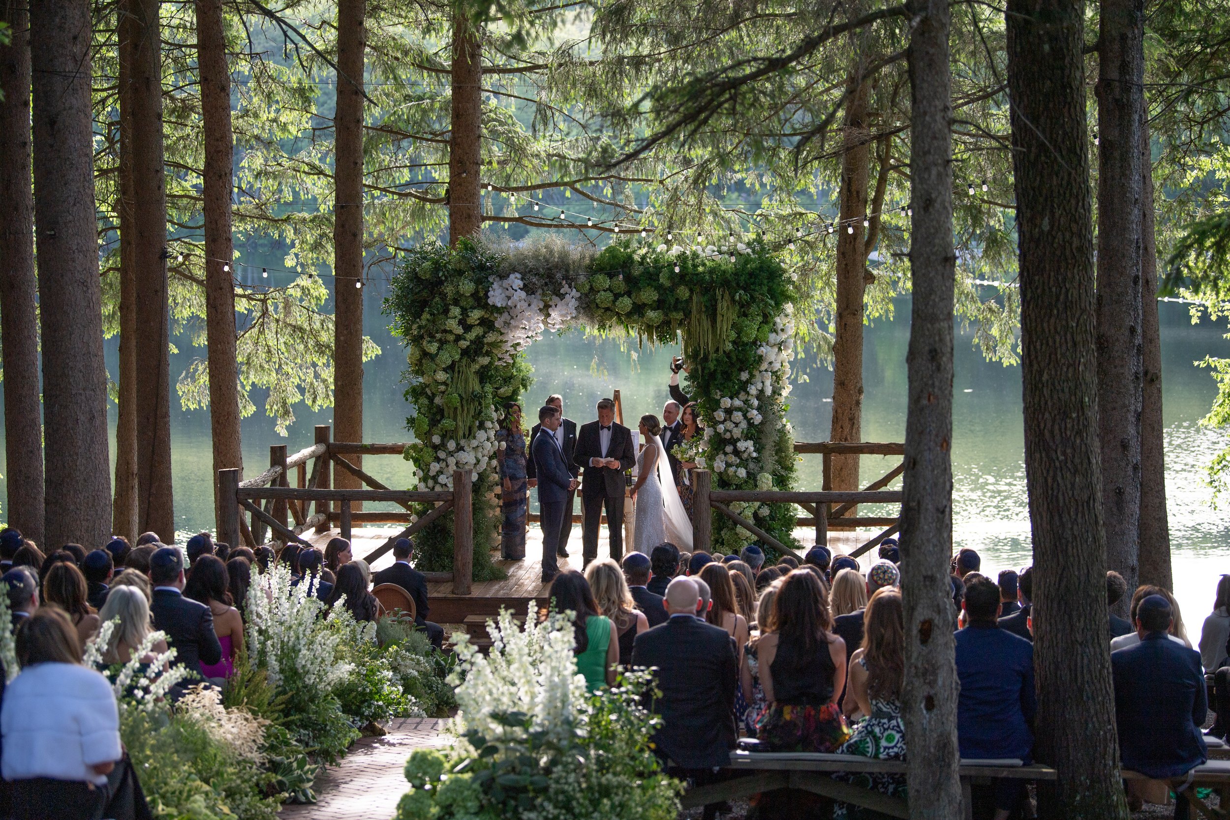 Outdoor wedding ceremony by a lake in a wooded area with a floral arch and string lights, surrounded by seated guests.