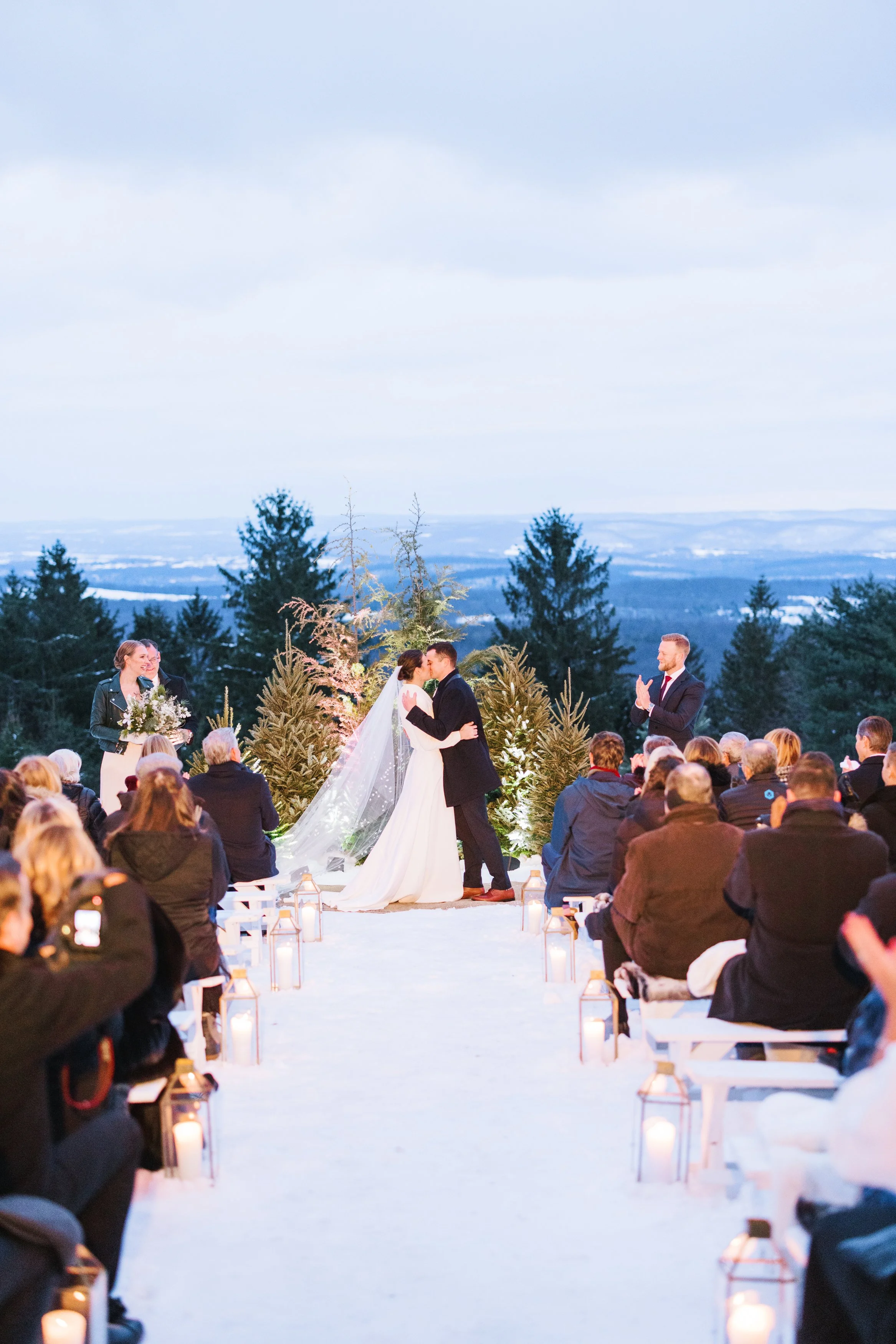 A winter outdoor wedding ceremony with a bride and groom kissing, surrounded by guests seated on white benches, with candle lanterns along the aisle and a backdrop of trees and snowy hills.