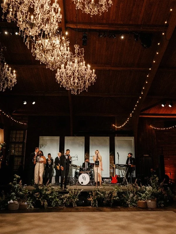 A live band performing on stage in a rustic venue with chandeliers and string lights, surrounded by floral arrangements.