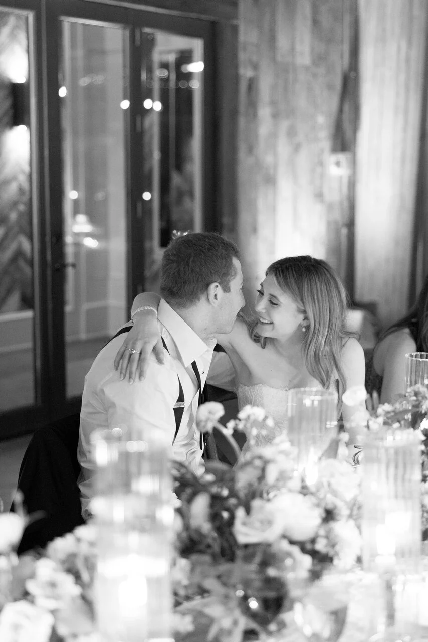A black and white photo of a couple smiling and embracing at a dinner table during a wedding reception, with floral centerpieces and candles.