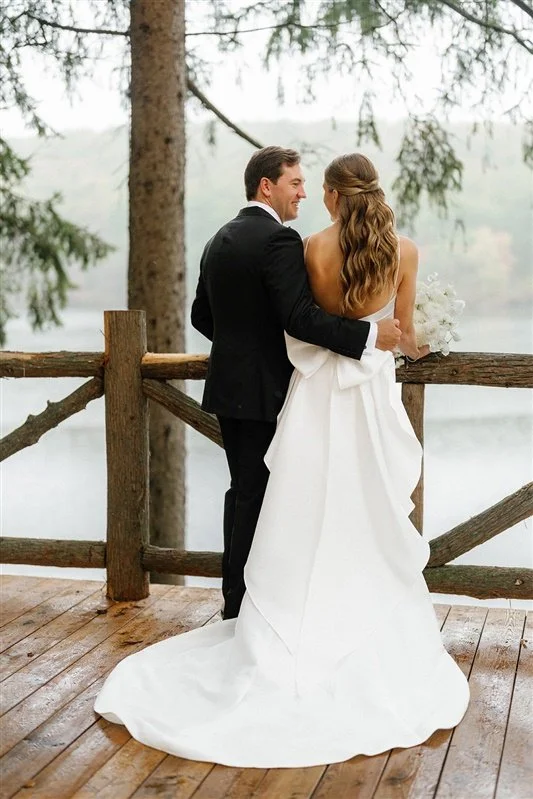 A newlywed couple standing on a wooden deck near a lake, embracing and smiling at each other, with trees and water in the background.
