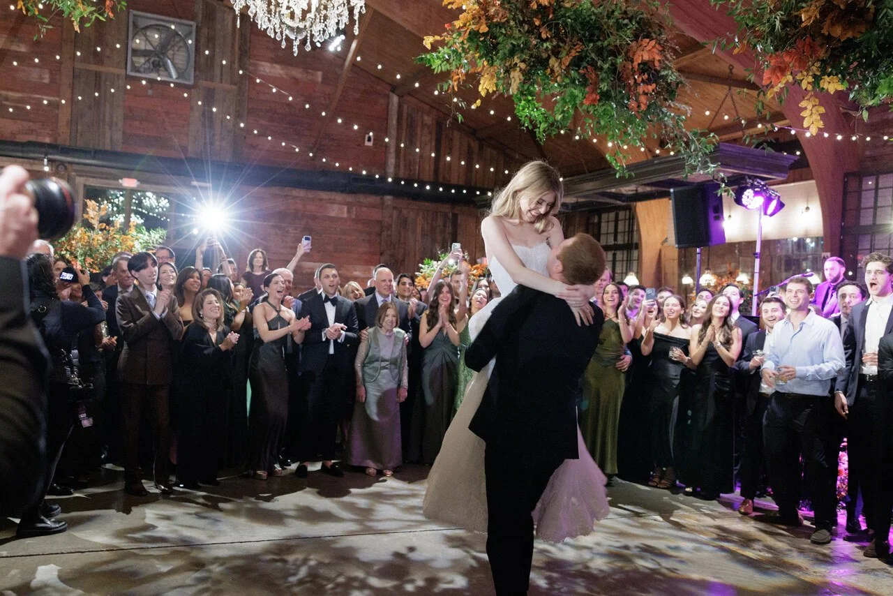 A bride and groom sharing a dance surrounded by wedding guests in a rustic indoor venue with wood-paneled walls, hanging lights, and floral decorations.