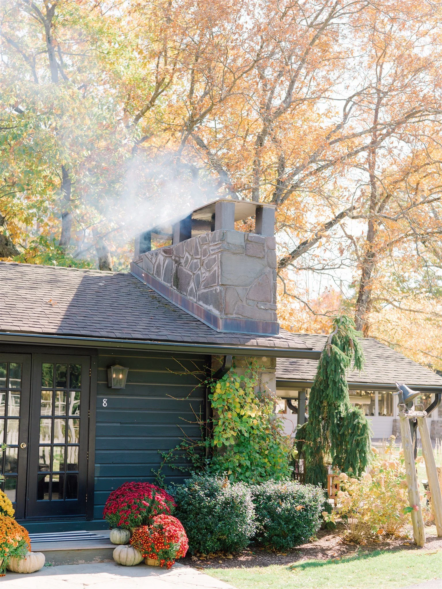 A house with green exterior walls, surrounded by colorful fall flowers and pumpkins, with a chimney emitting smoke, set against a backdrop of orange and yellow autumn trees.