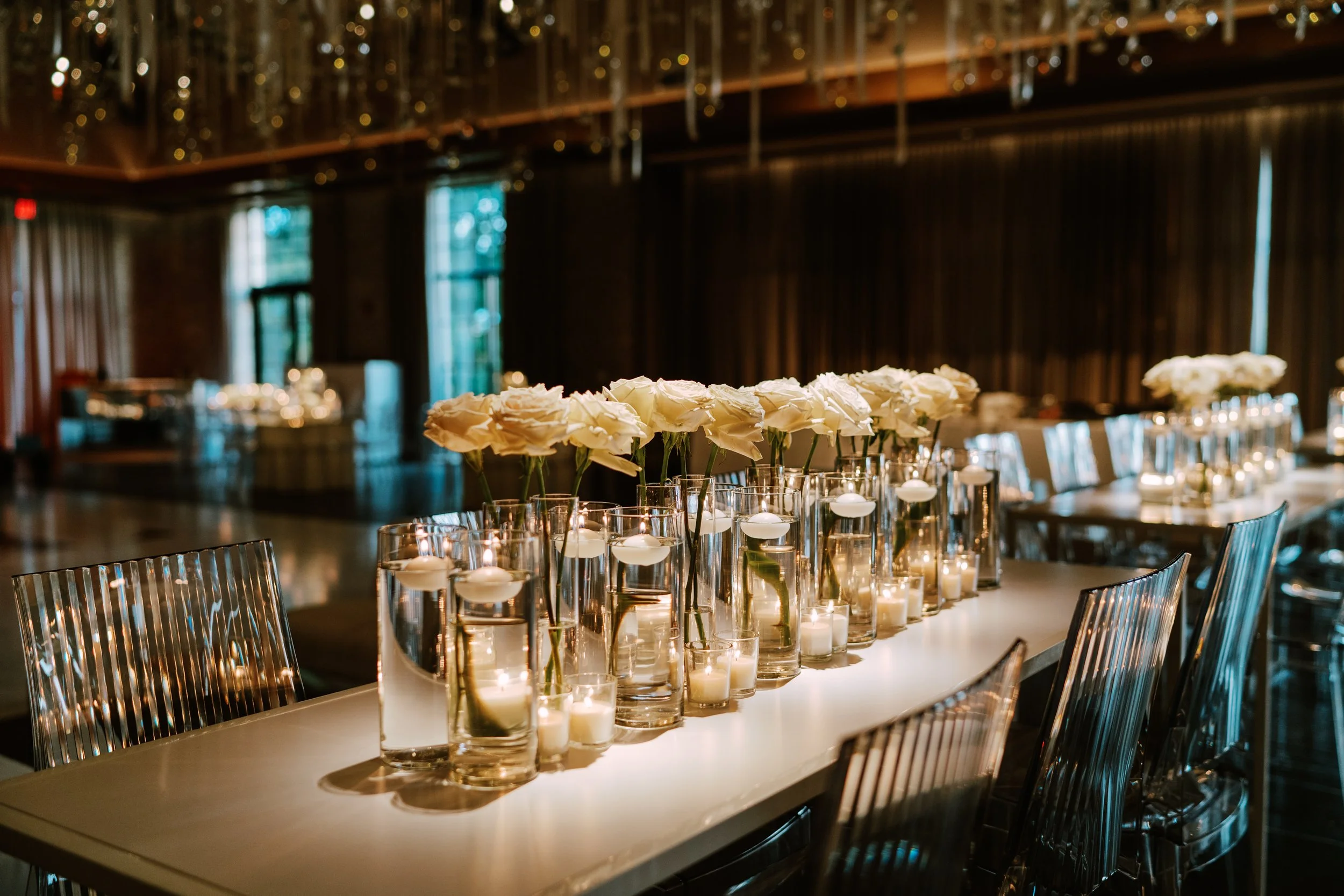 Elegant indoor event space with a long table decorated with white roses in glass vases and small floating candles, set against a backdrop of dark wood paneling and hanging glass decor.