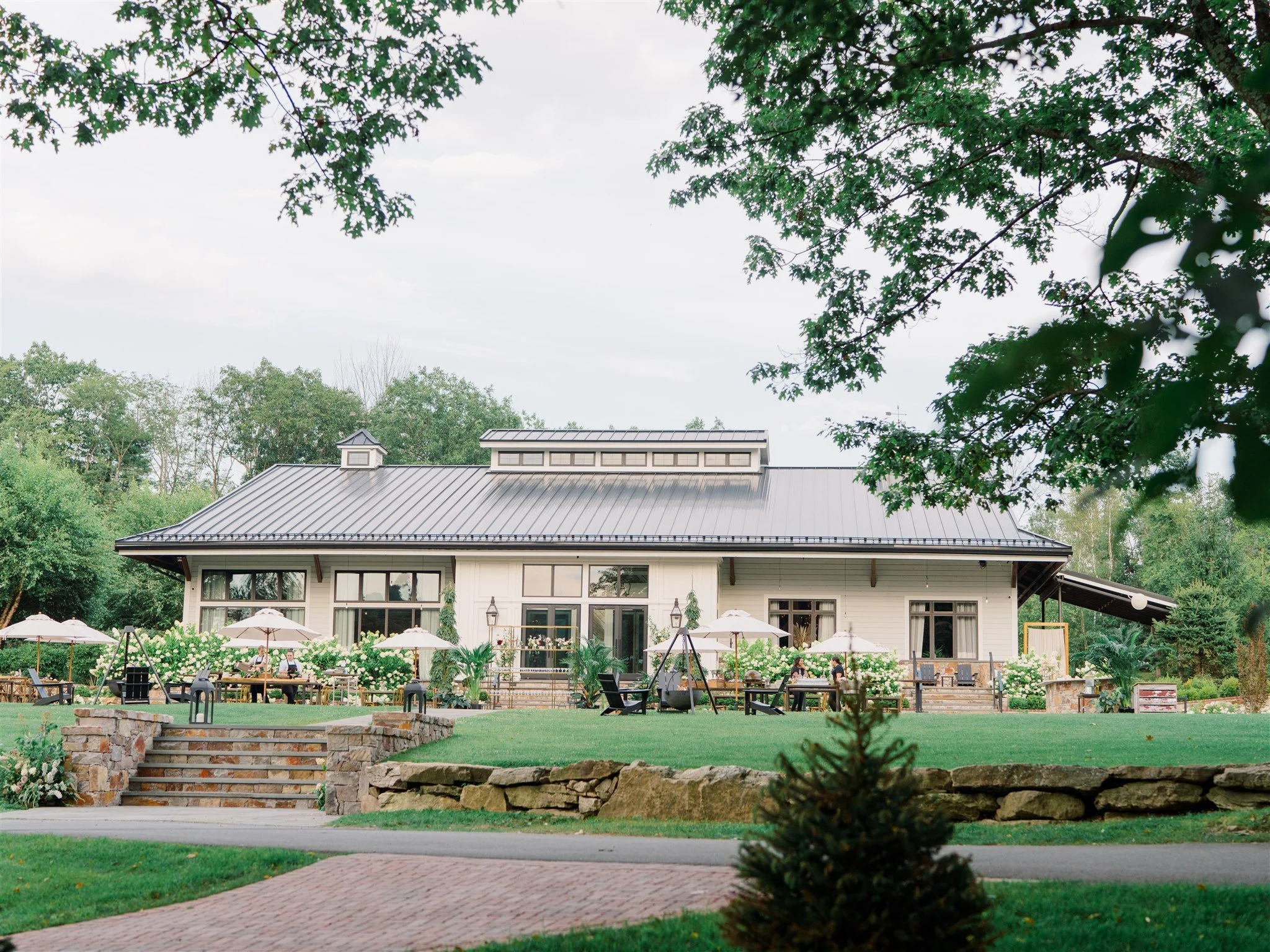A large house with a metal roof and multiple windows, surrounded by a well-maintained lawn with outdoor patio furniture and umbrellas, set in a lush green environment with trees.