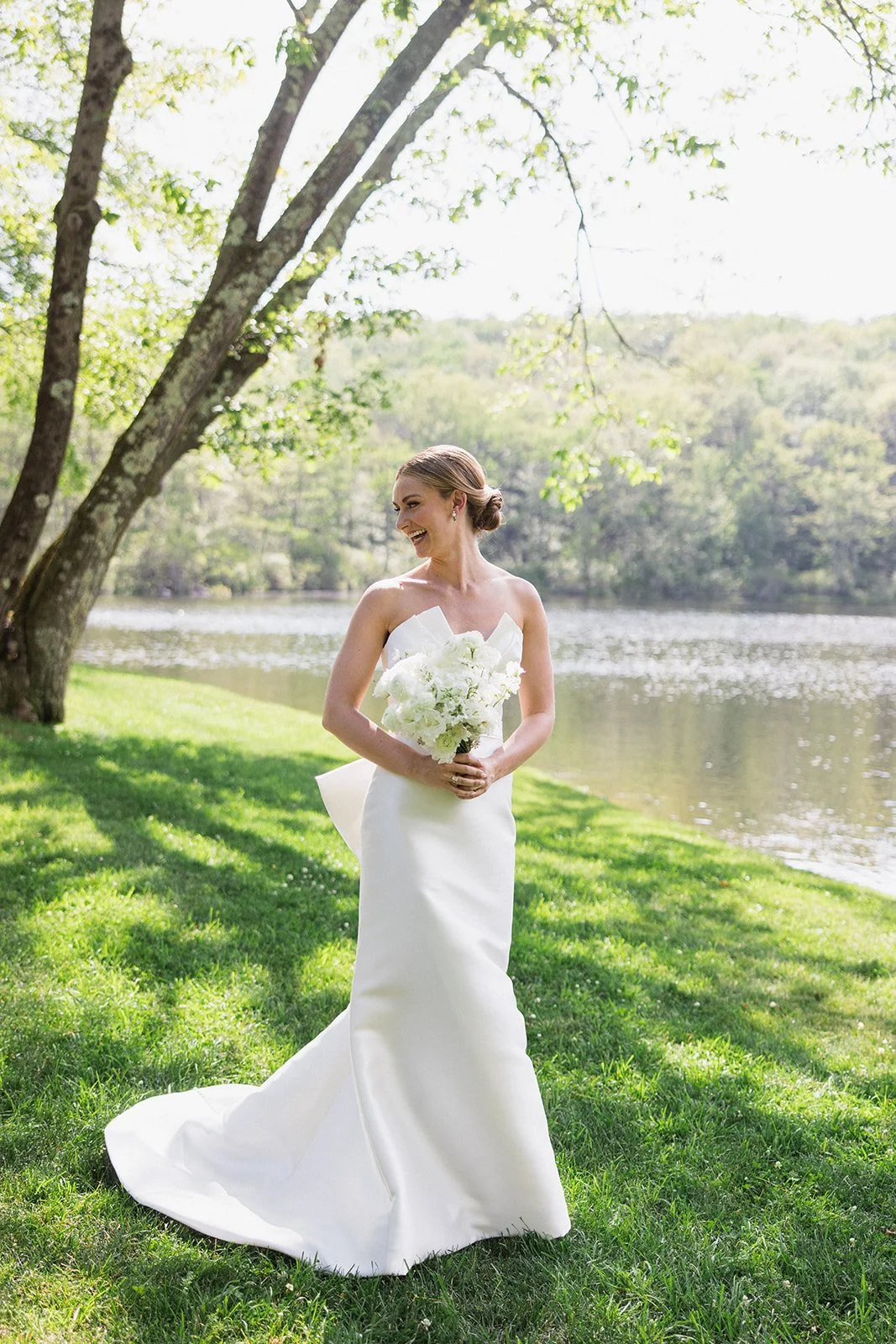 A bride in a white wedding dress holding a bouquet of white flowers outdoors near a lake, with green grass and trees in the background.