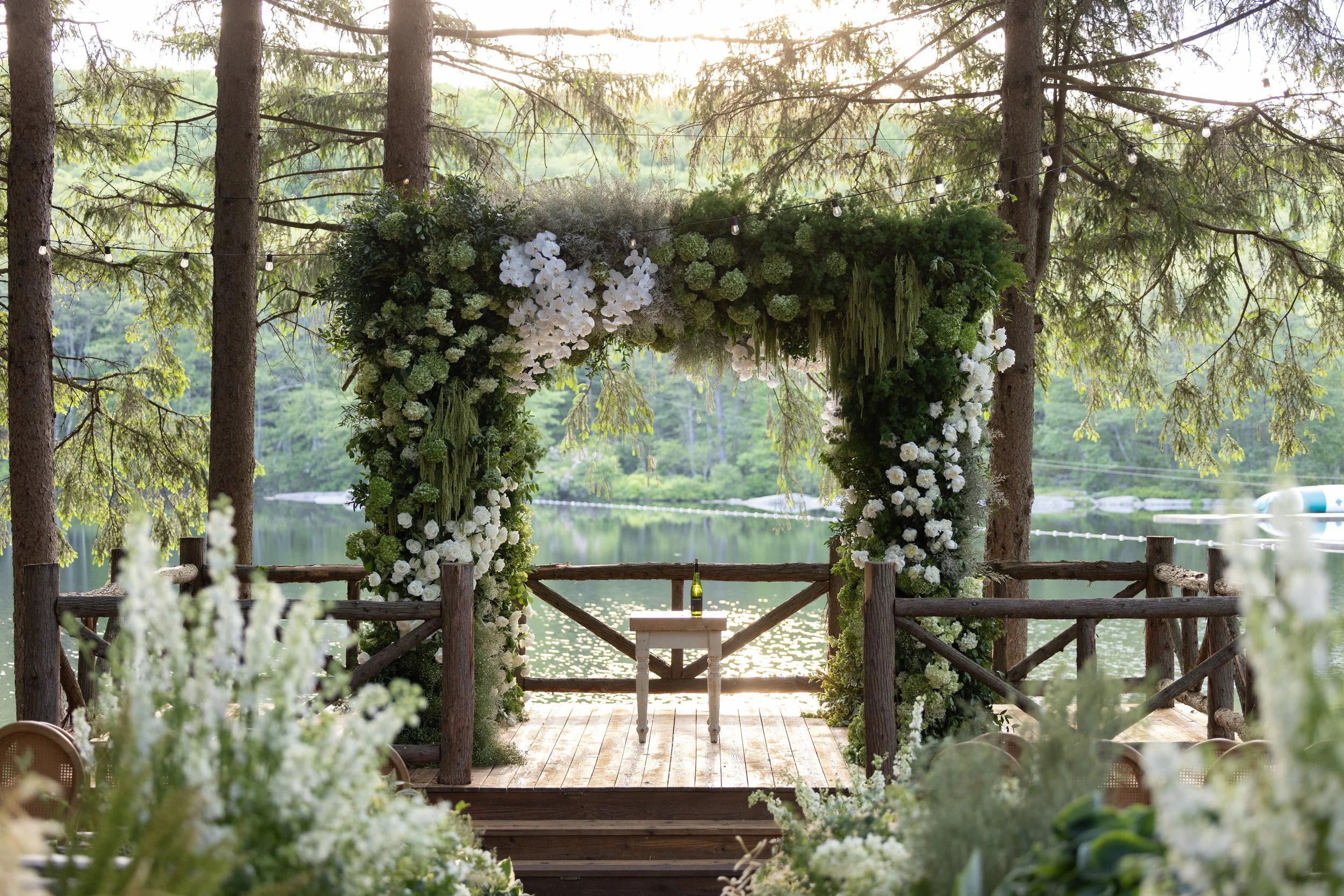 A decorated outdoor wedding arch with white flowers in front of a lake surrounded by trees, with a small table and a bottle of drink underneath the arch.